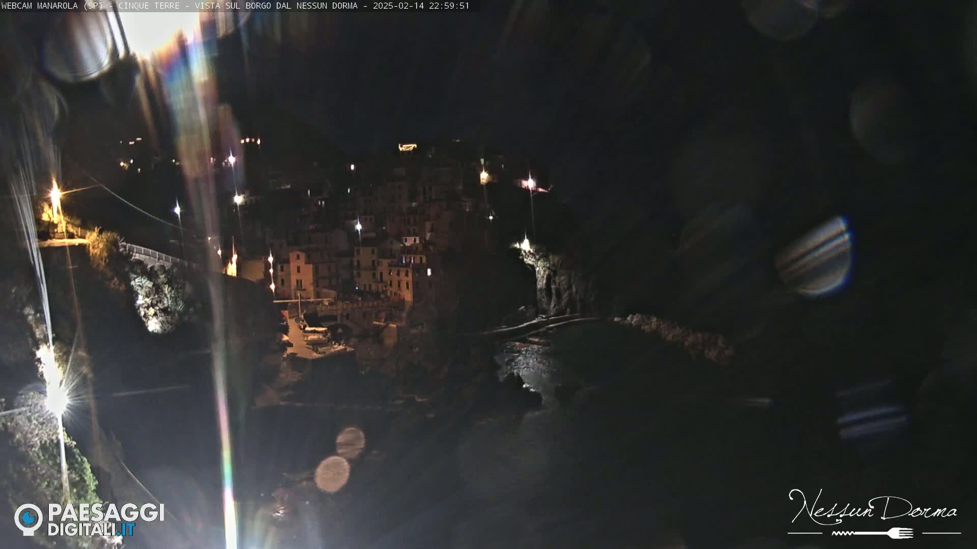 A nighttime view of a coastal town nestled between dark cliffs, illuminated by scattered lights.