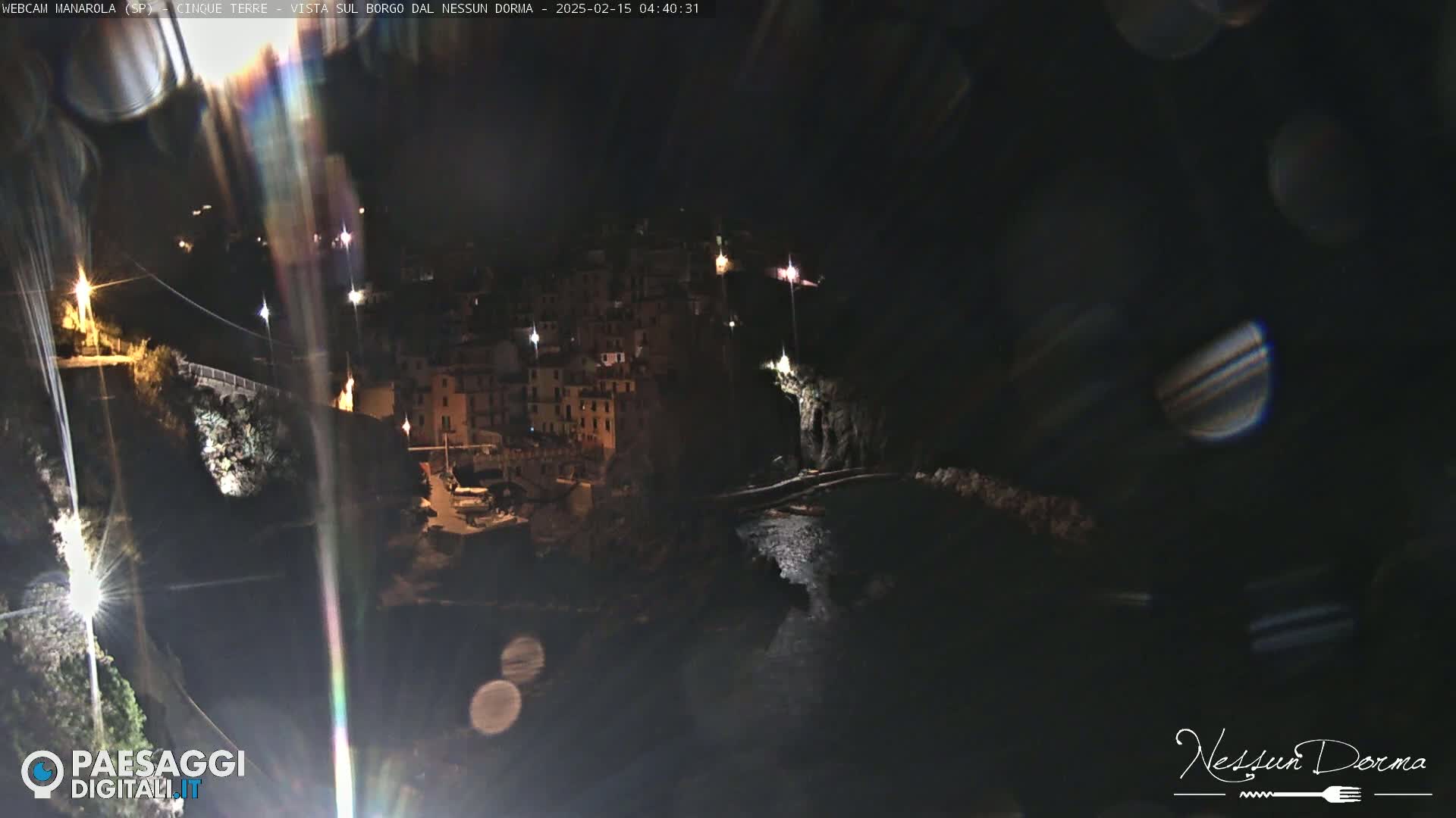 A nighttime, coastal village with buildings illuminated clings to a dark cliffside overlooking a calm body of water.