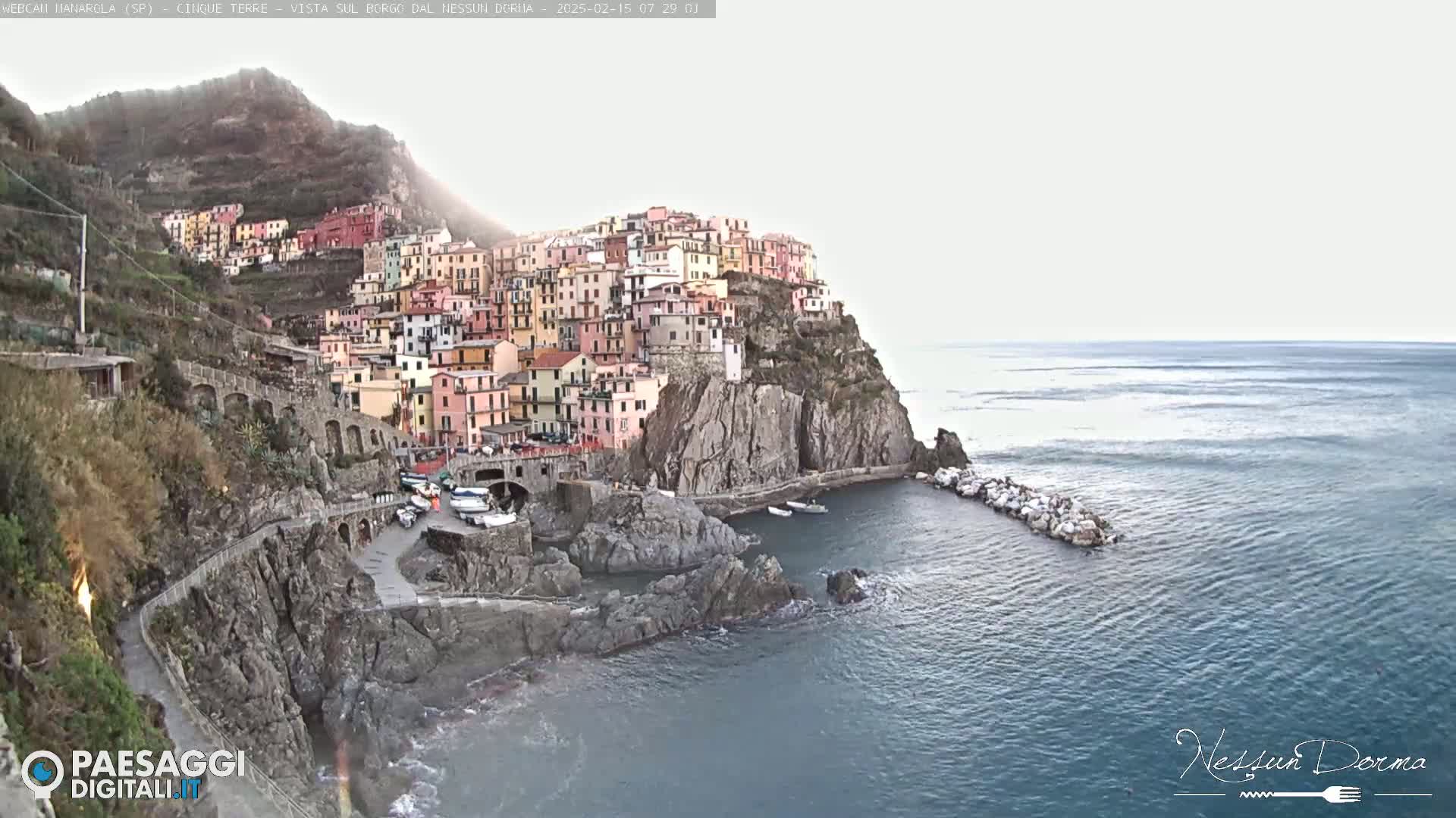 A colorful village clings to a rocky cliffside overlooking a calm sea under a mostly clear sky.