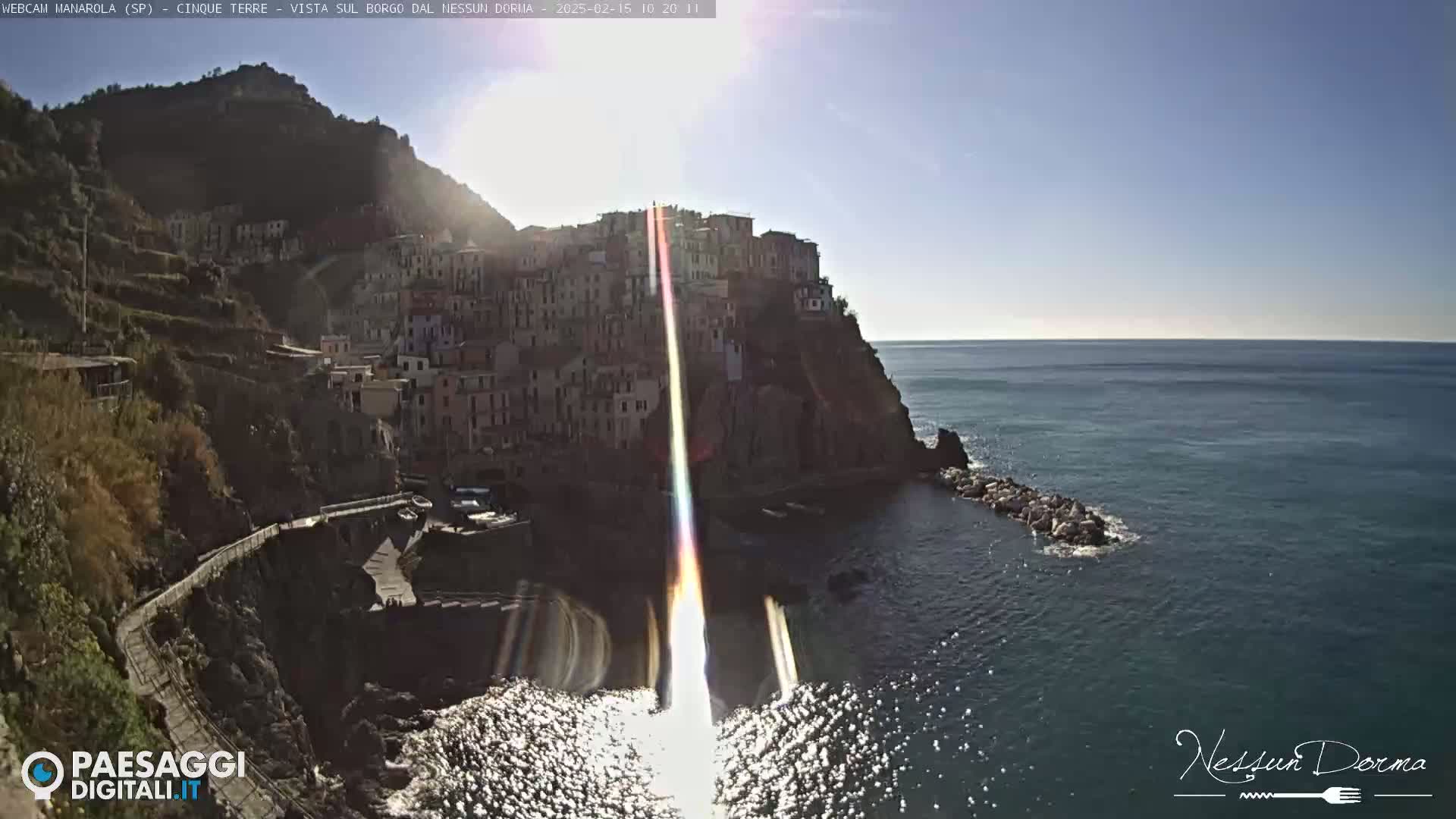 A sunlit coastal town clings to a cliffside overlooking a calm, blue sea on a bright, clear day.