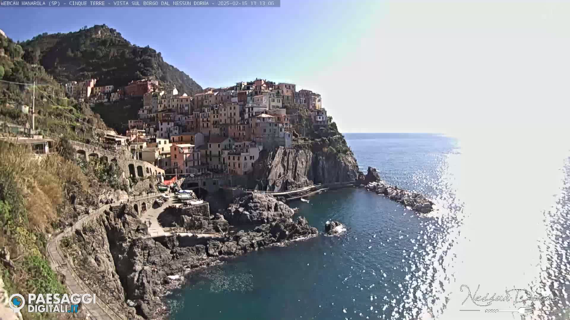 A colorful village clings to a rocky cliffside overlooking a calm, blue sea under a bright, sunny sky.
