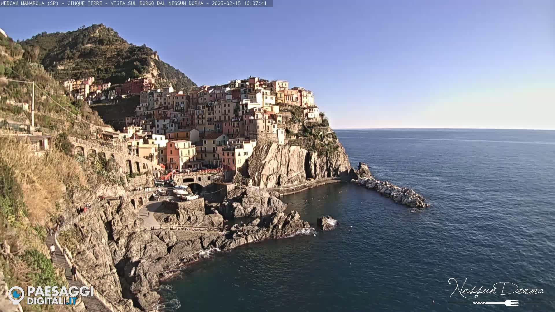 A picturesque coastal village clings to a rocky cliffside overlooking a calm sea under a clear, sunny sky.