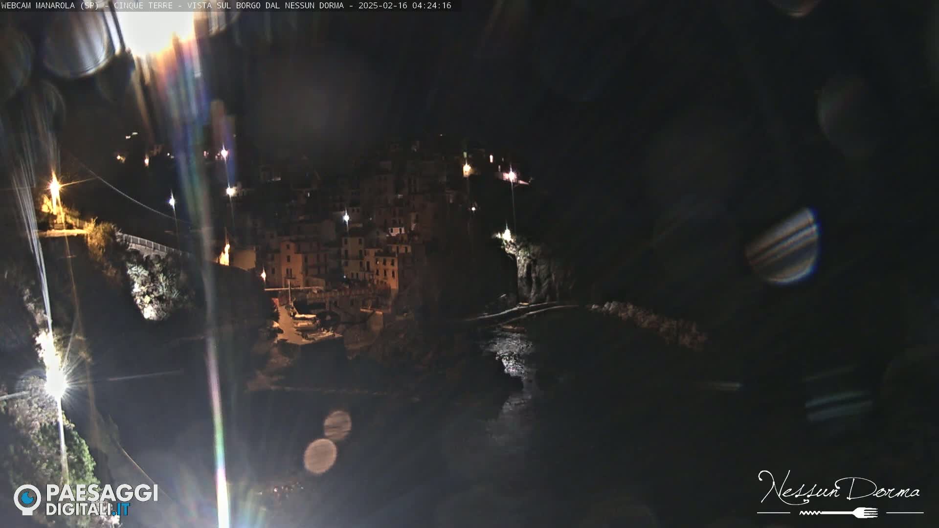 A nighttime, coastal village is illuminated by scattered lights, nestled between dark cliffs and a body of water.