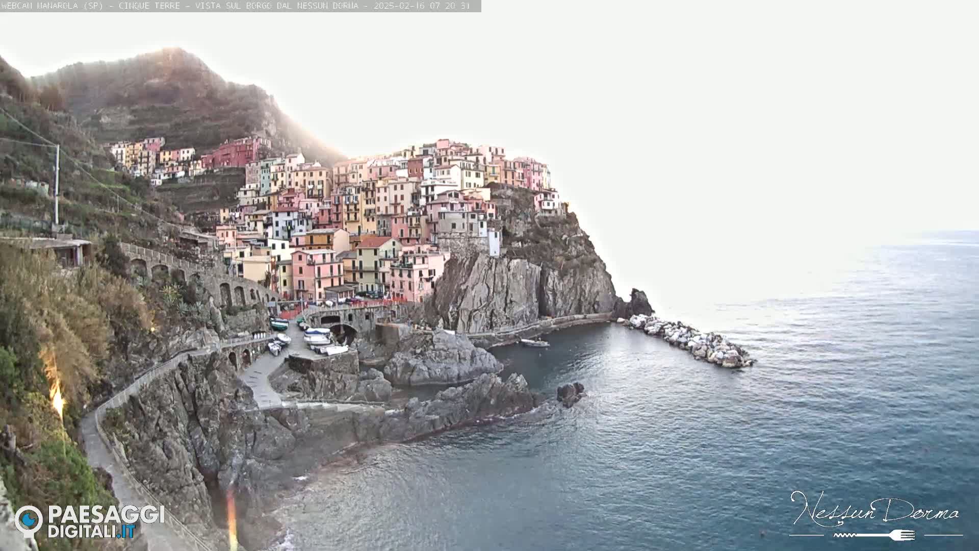 A colorful village clings to a rocky cliffside overlooking a calm sea under a hazy sky.