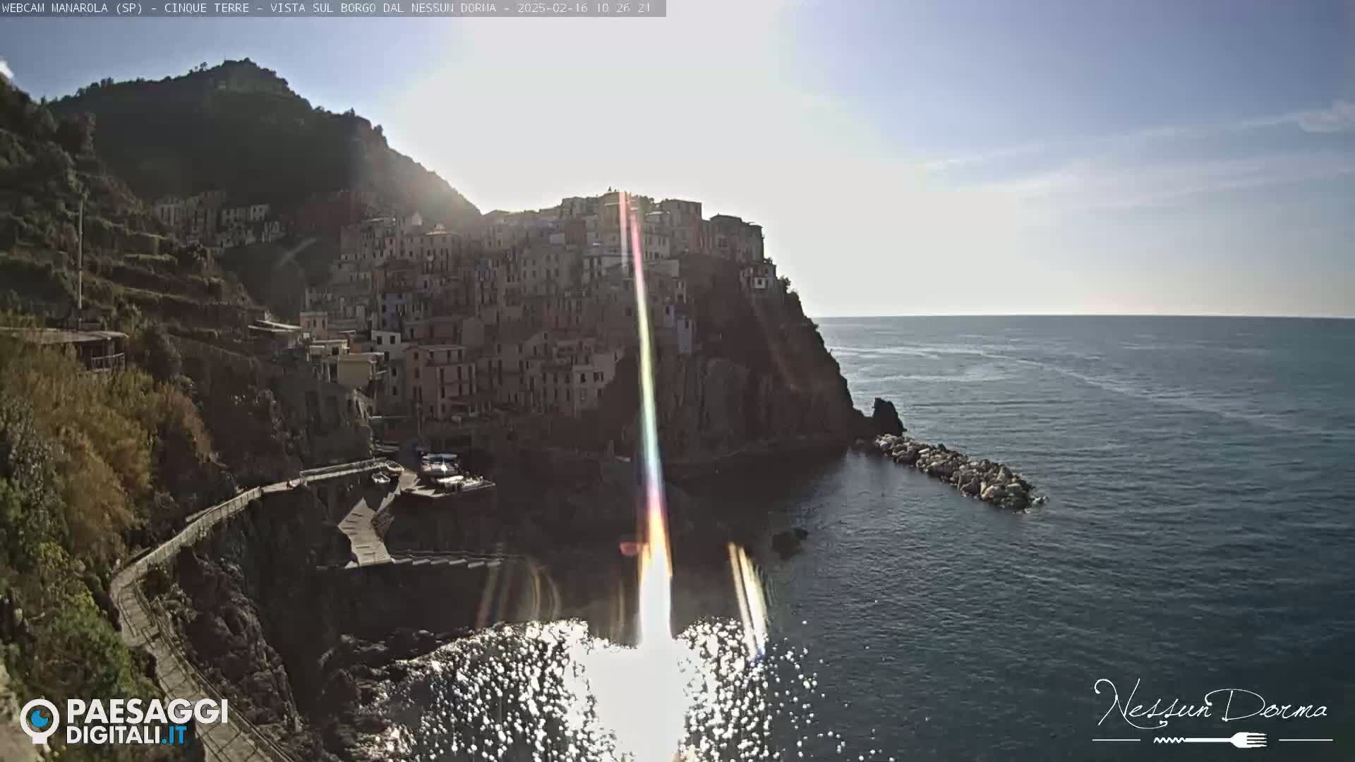 A coastal village clings to a steep cliffside overlooking a calm sea under a bright, sunny sky.