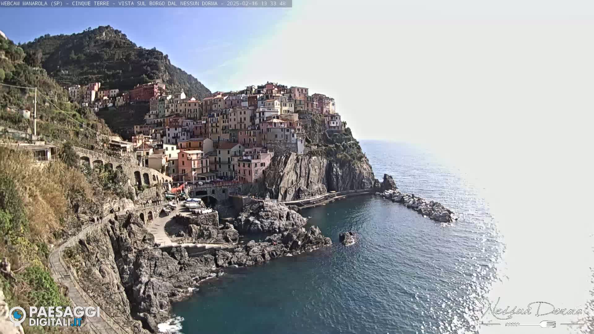 A colorful village clings to a steep cliffside overlooking a calm sea under a bright, clear sky.