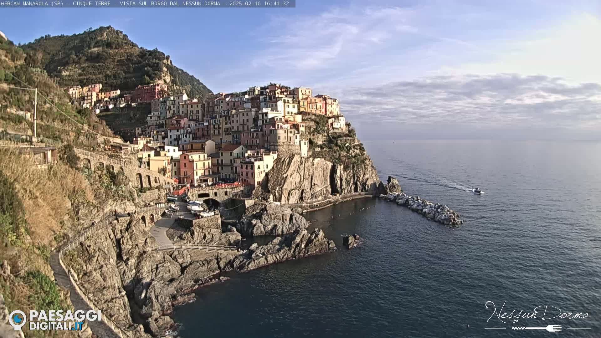A picturesque coastal village clings to a rocky cliffside overlooking a calm sea under a partly cloudy sky.