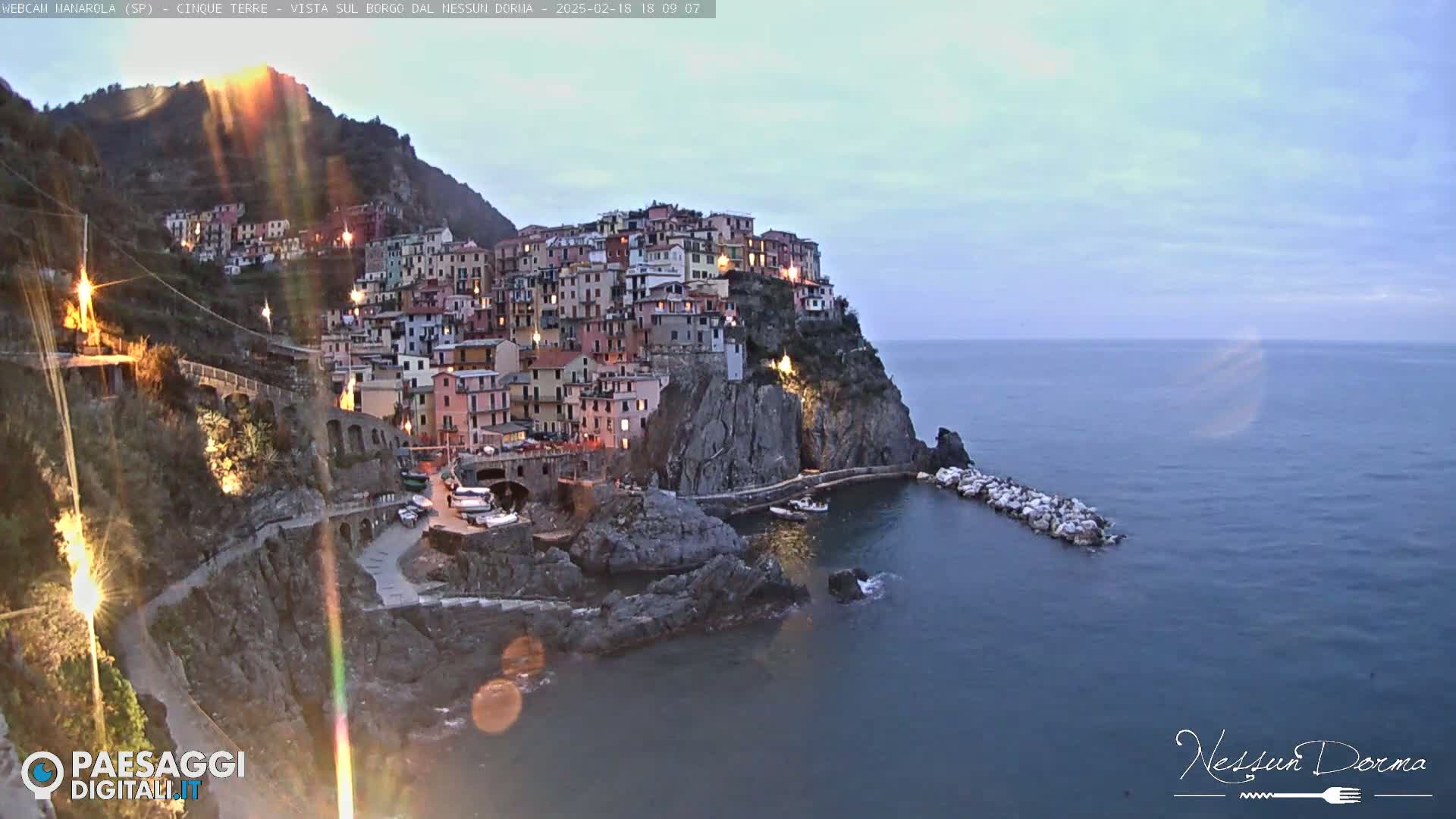 A brightly lit coastal village clings to a steep cliffside overlooking a calm, dark blue sea under a twilight sky.