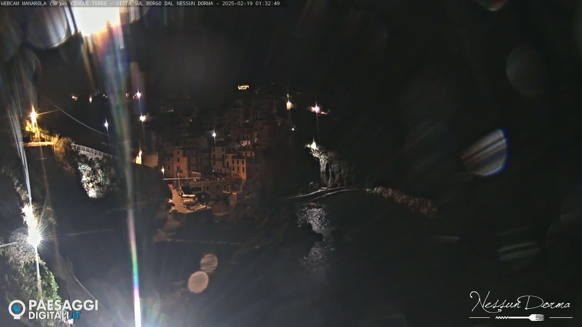 A nighttime view of a coastal town, illuminated by scattered lights, with dark water visible below.