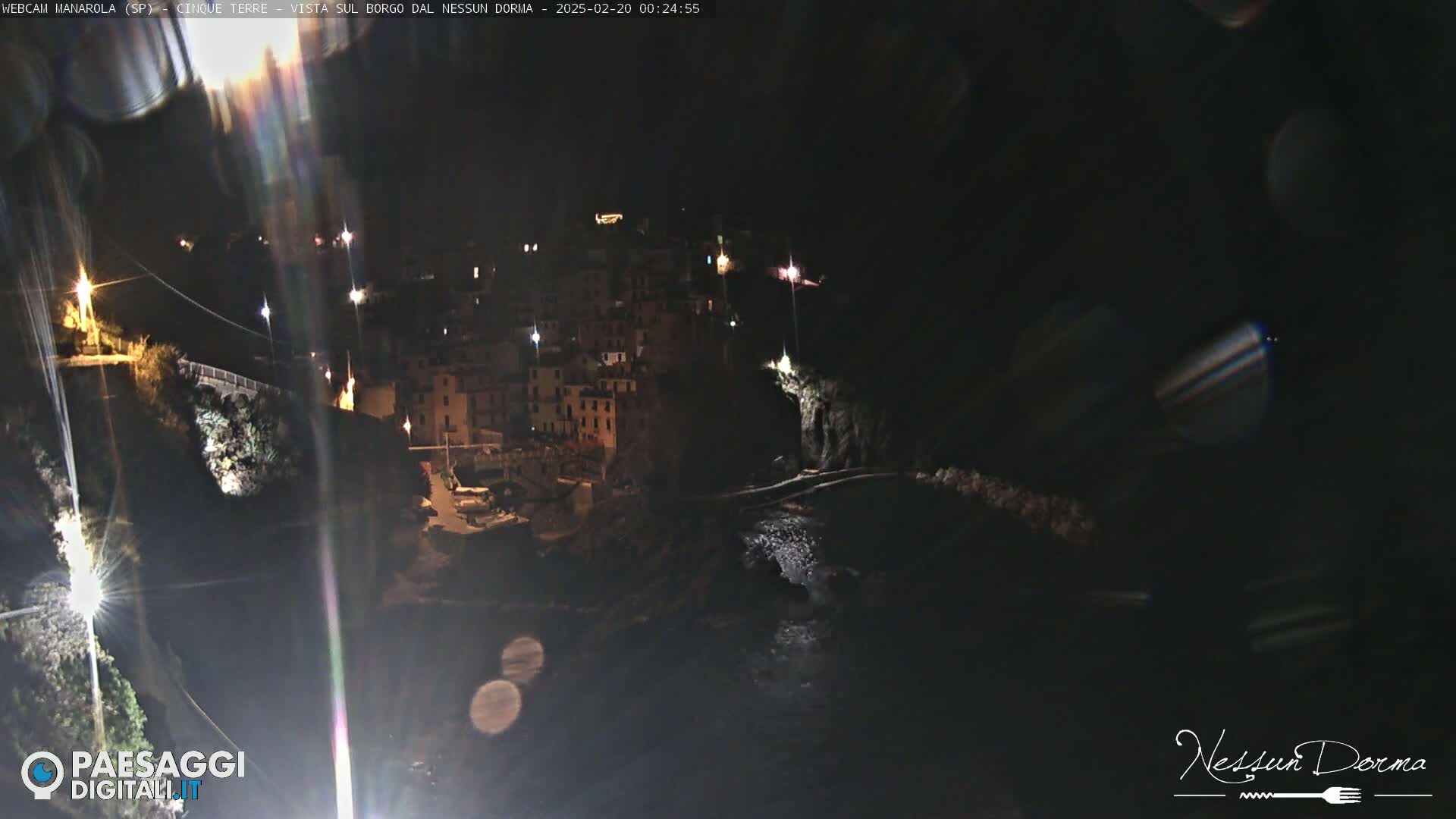 A nighttime, long shot of a coastal town with buildings illuminated on a dark cliffside overlooking a body of water.