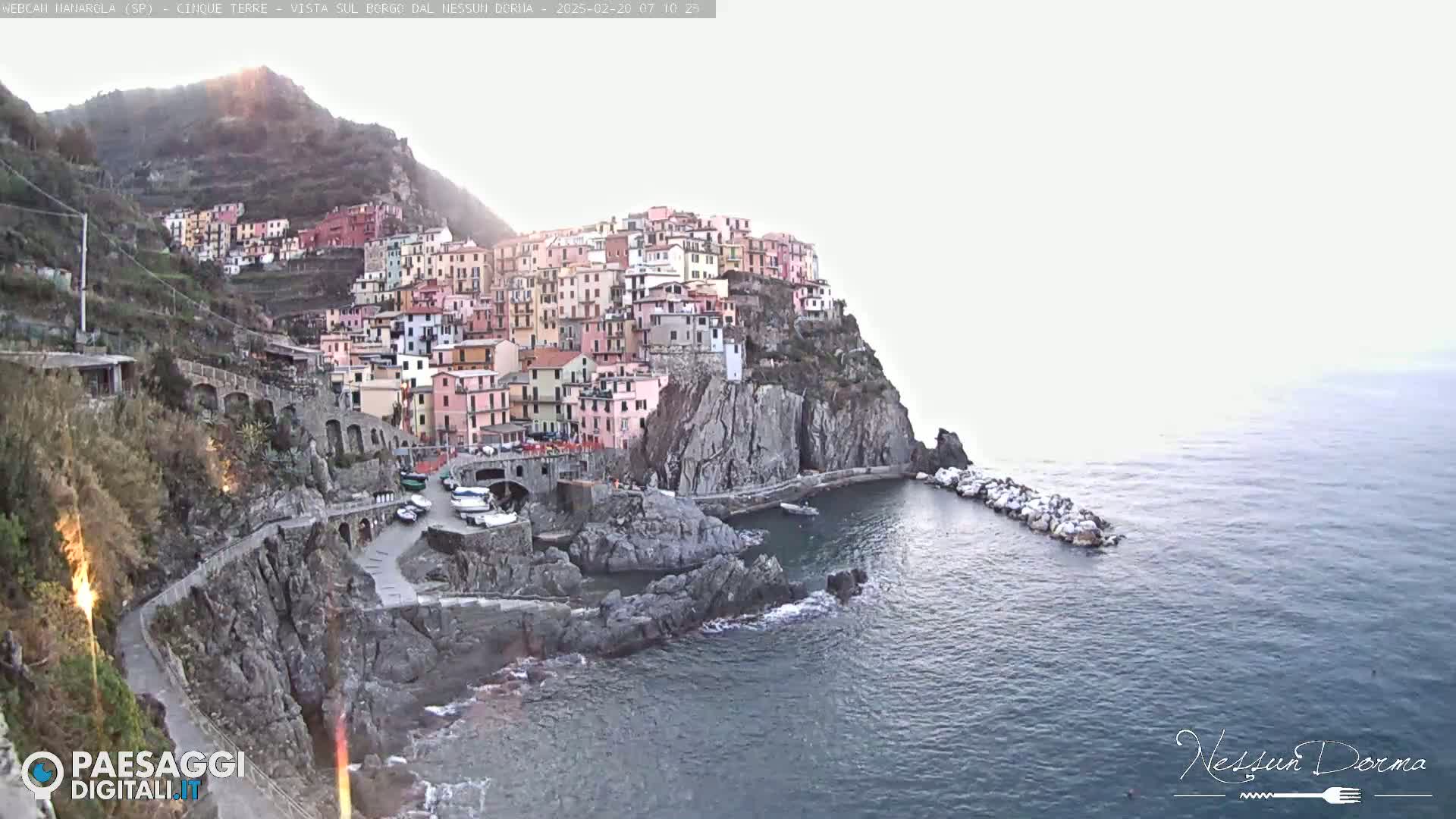 A colorful village clings to a steep cliffside overlooking a calm sea under a hazy sky.