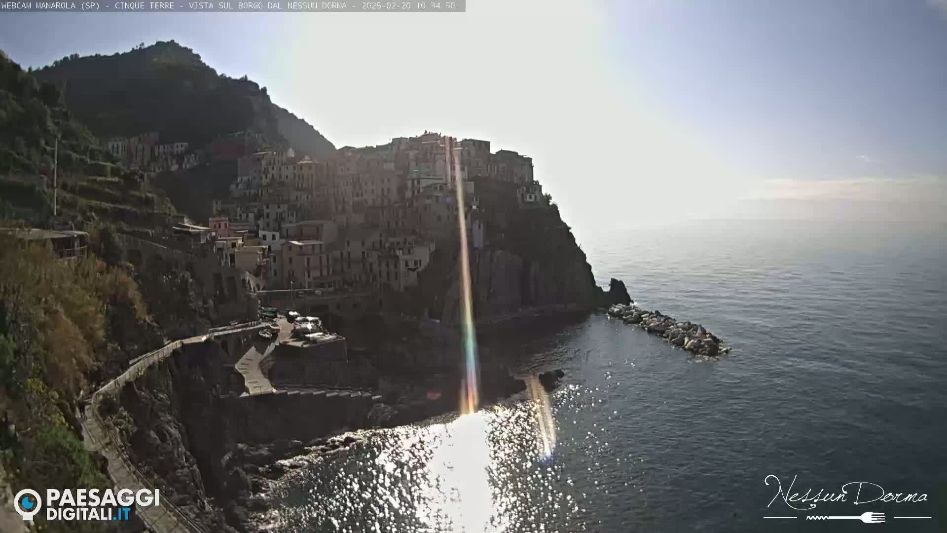 A sunlit coastal village clings to a steep cliffside overlooking a calm sea.