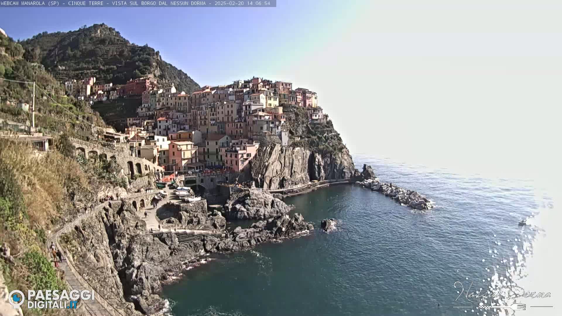 A colorful village clings to a steep cliffside overlooking a calm sea under a bright, sunny sky.