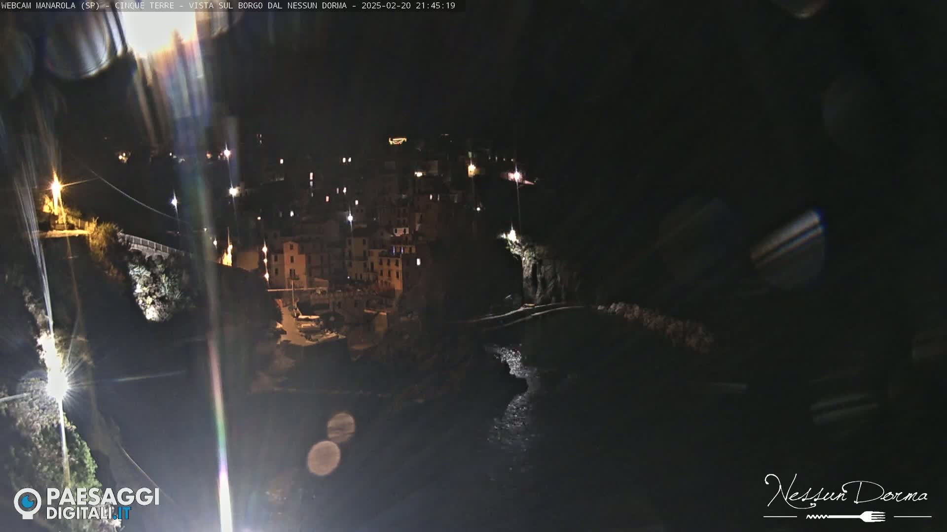 A nighttime view of a coastal village with buildings illuminated along a dark, rocky shoreline.