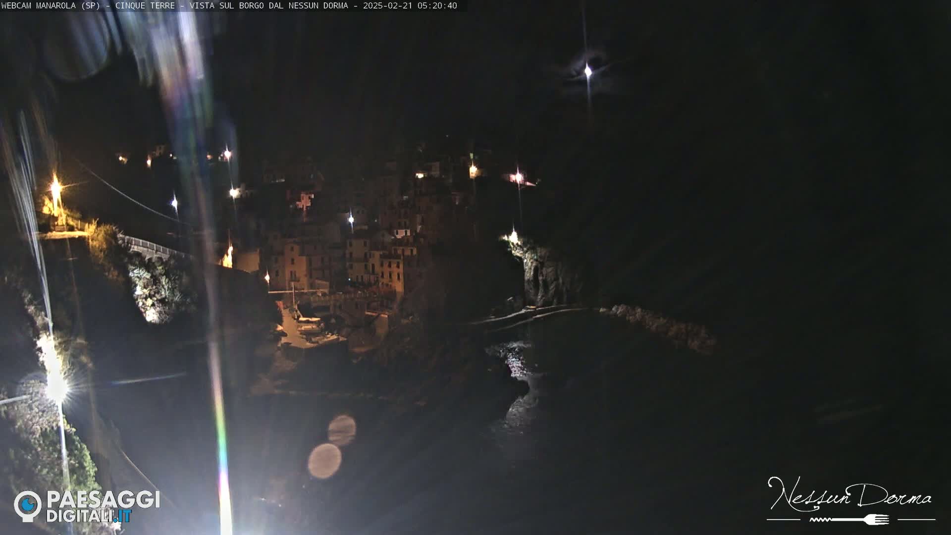 A nighttime, seaside village clings to a cliffside, illuminated by scattered lights, under a dark sky.