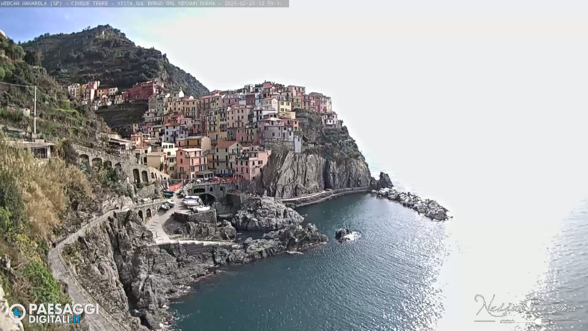A colorful village clings to a steep, rocky cliffside overlooking a calm, dark-blue sea under a mostly clear sky.