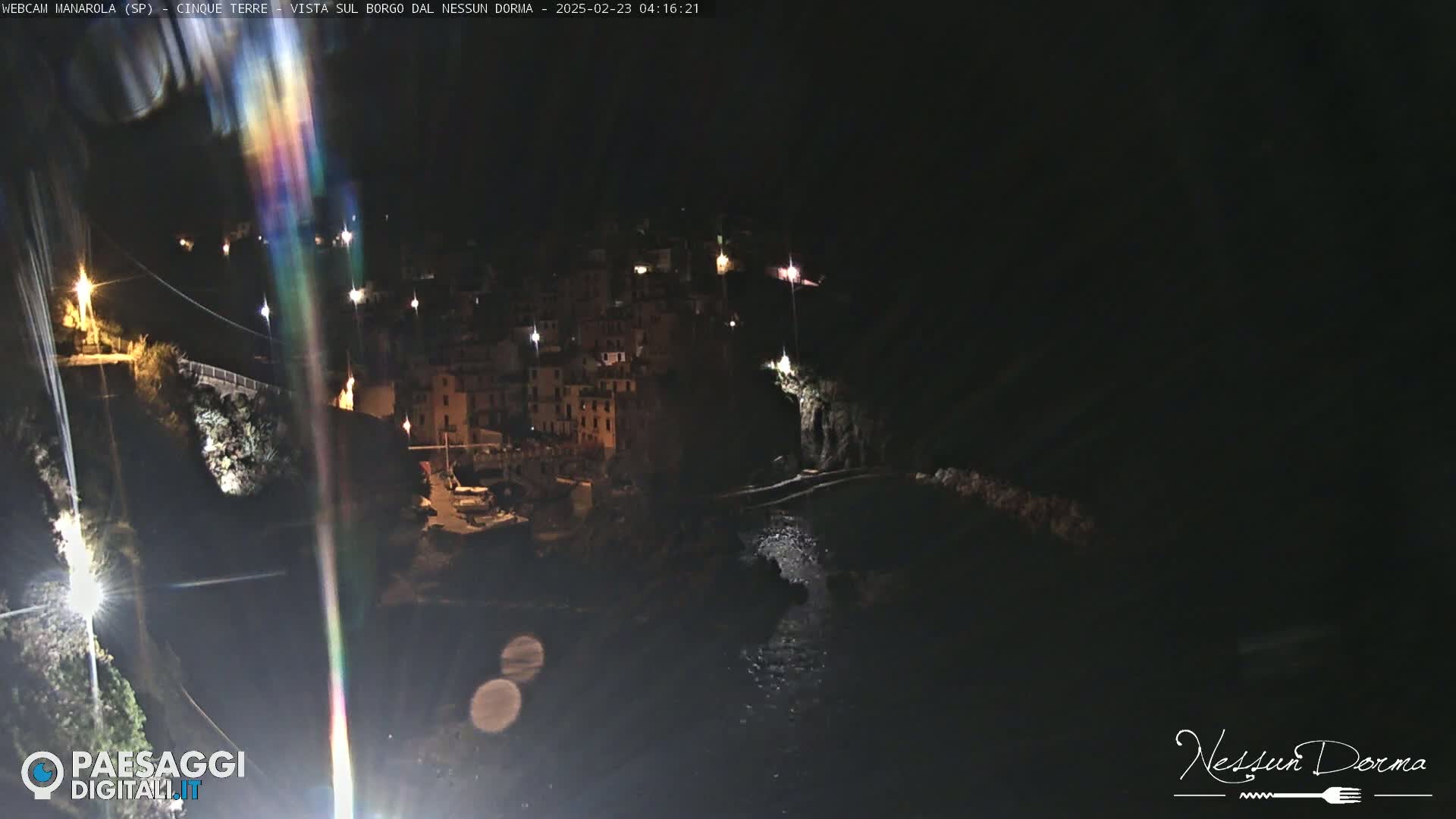 A nighttime, high-angle view of a coastal town illuminated by lights, with a dark body of water below.