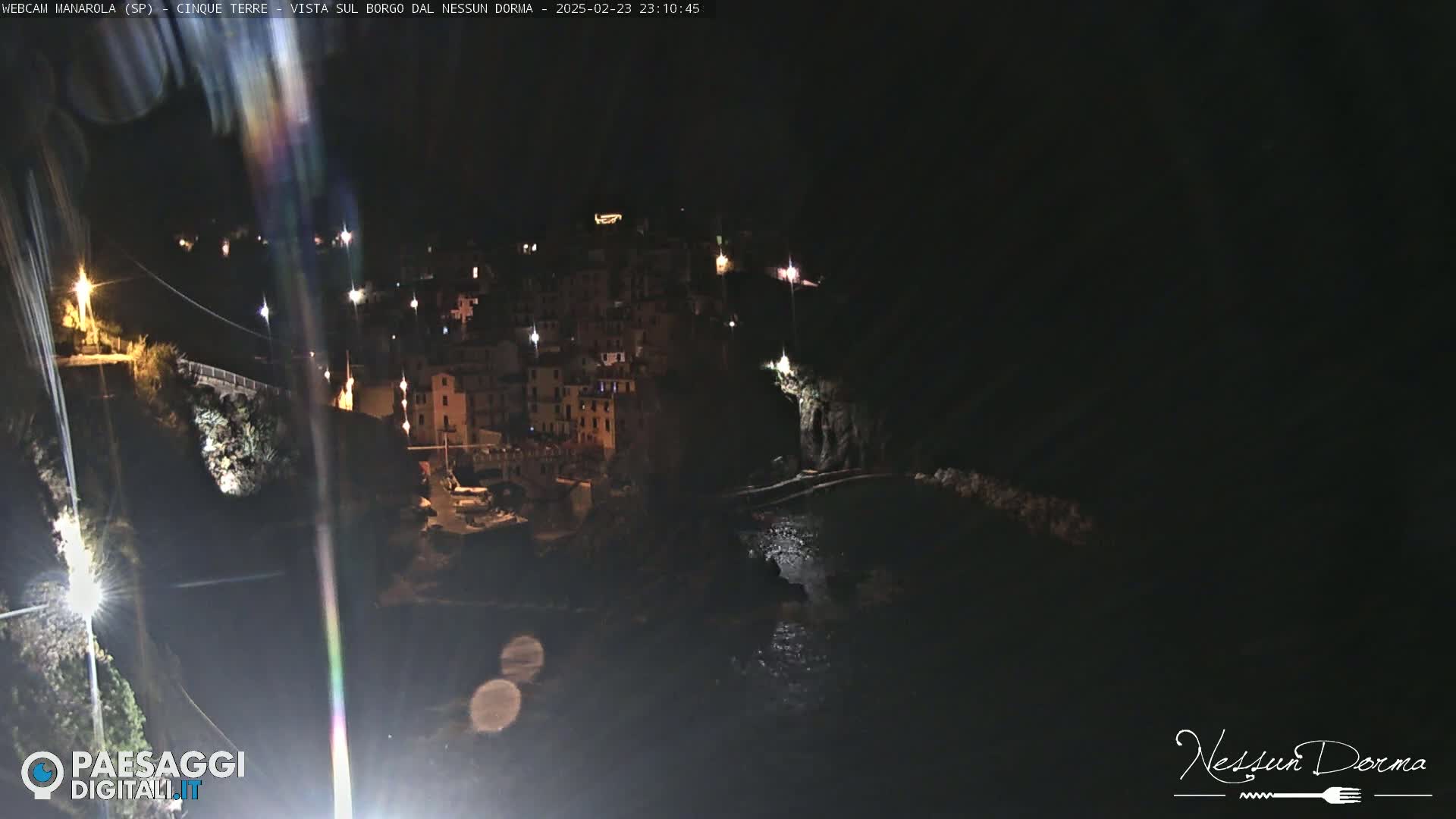 A nighttime view of a coastal town, illuminated by lights, with dark water below.