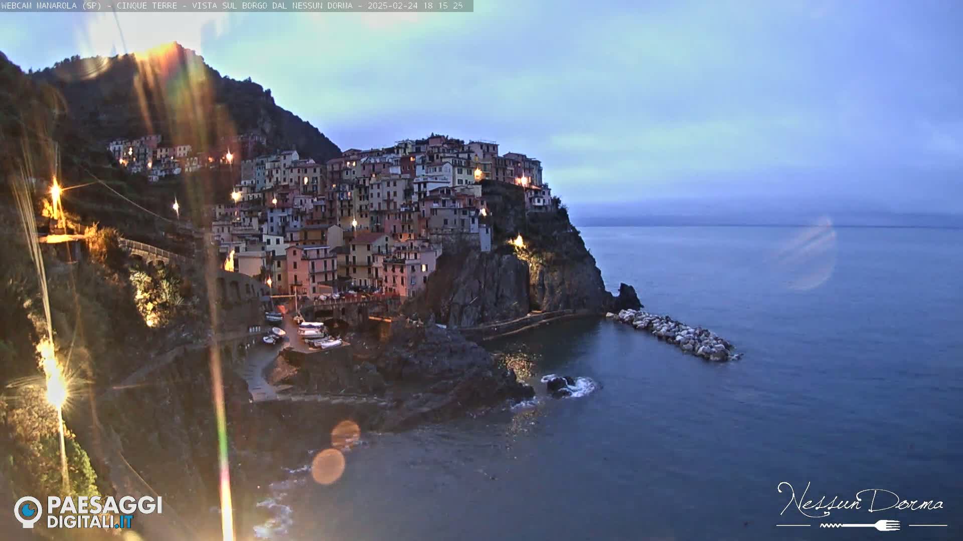 A coastal village clinging to a cliffside at twilight, illuminated by lights, under a mostly clear sky.