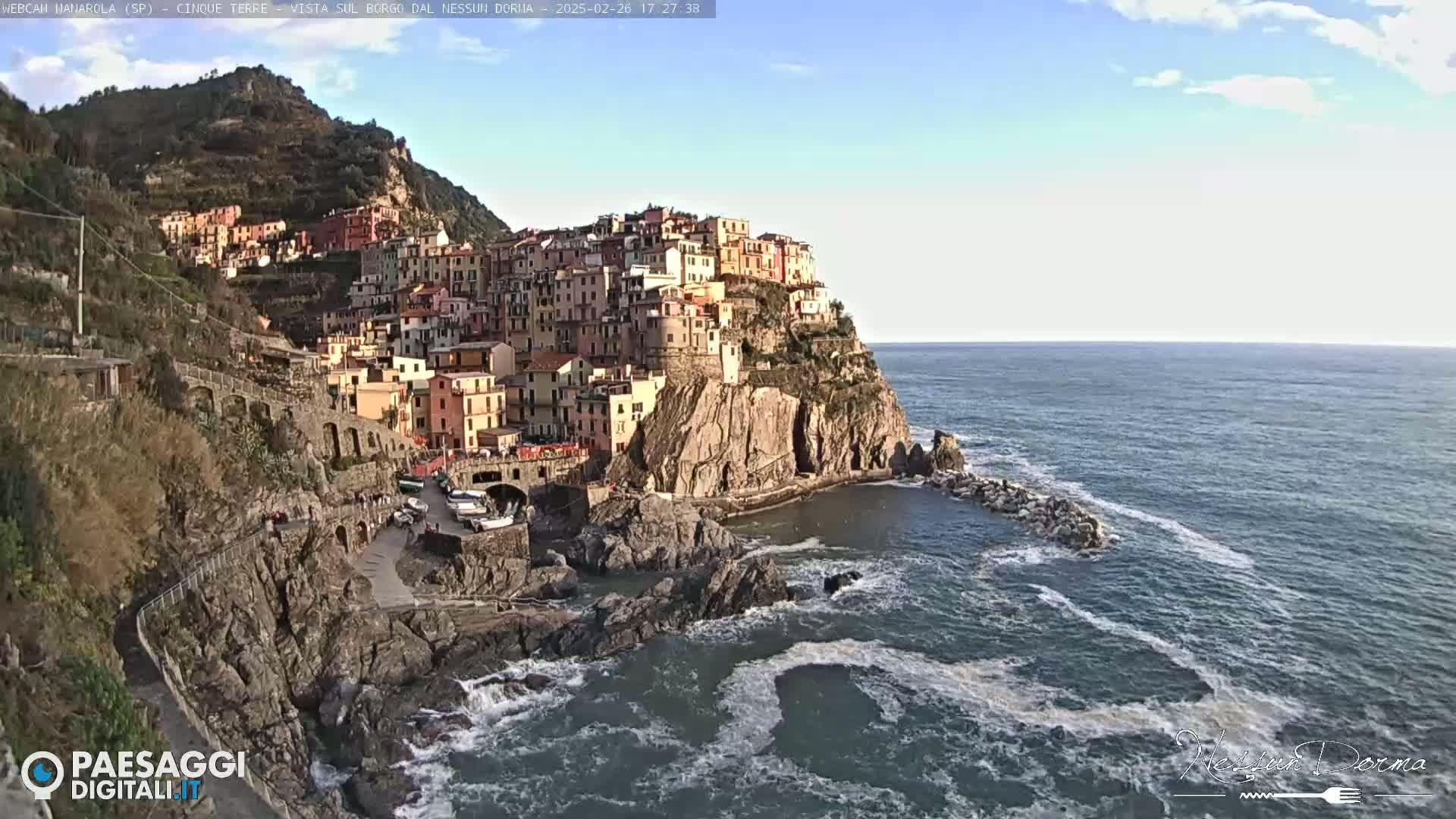A colorful village clings to a rocky cliffside overlooking a sea with moderate waves under a mostly clear sky.
