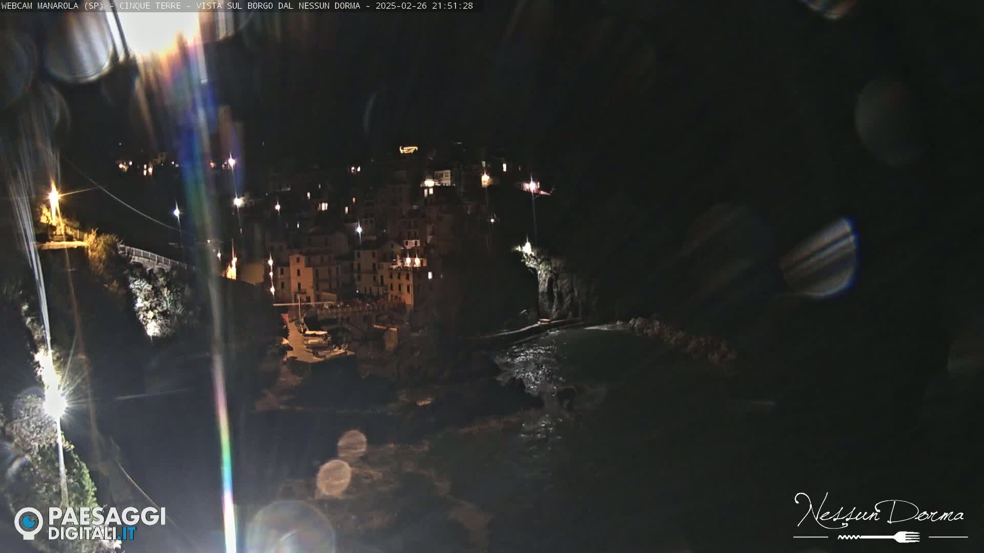 A nighttime view of a coastal town, illuminated by lights, with dark water lapping against rocky shores.