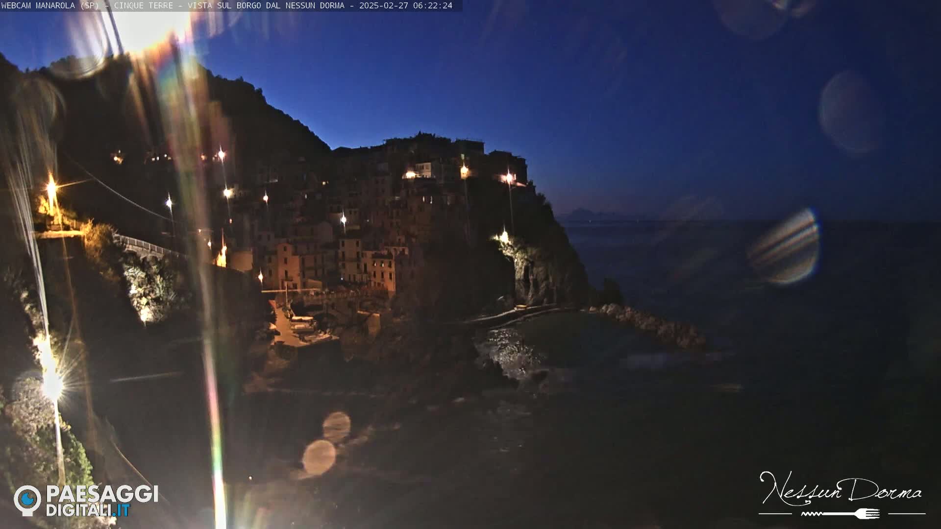 A nighttime view of a coastal town illuminated by lights, nestled on a cliff overlooking a dark ocean under a twilight sky.