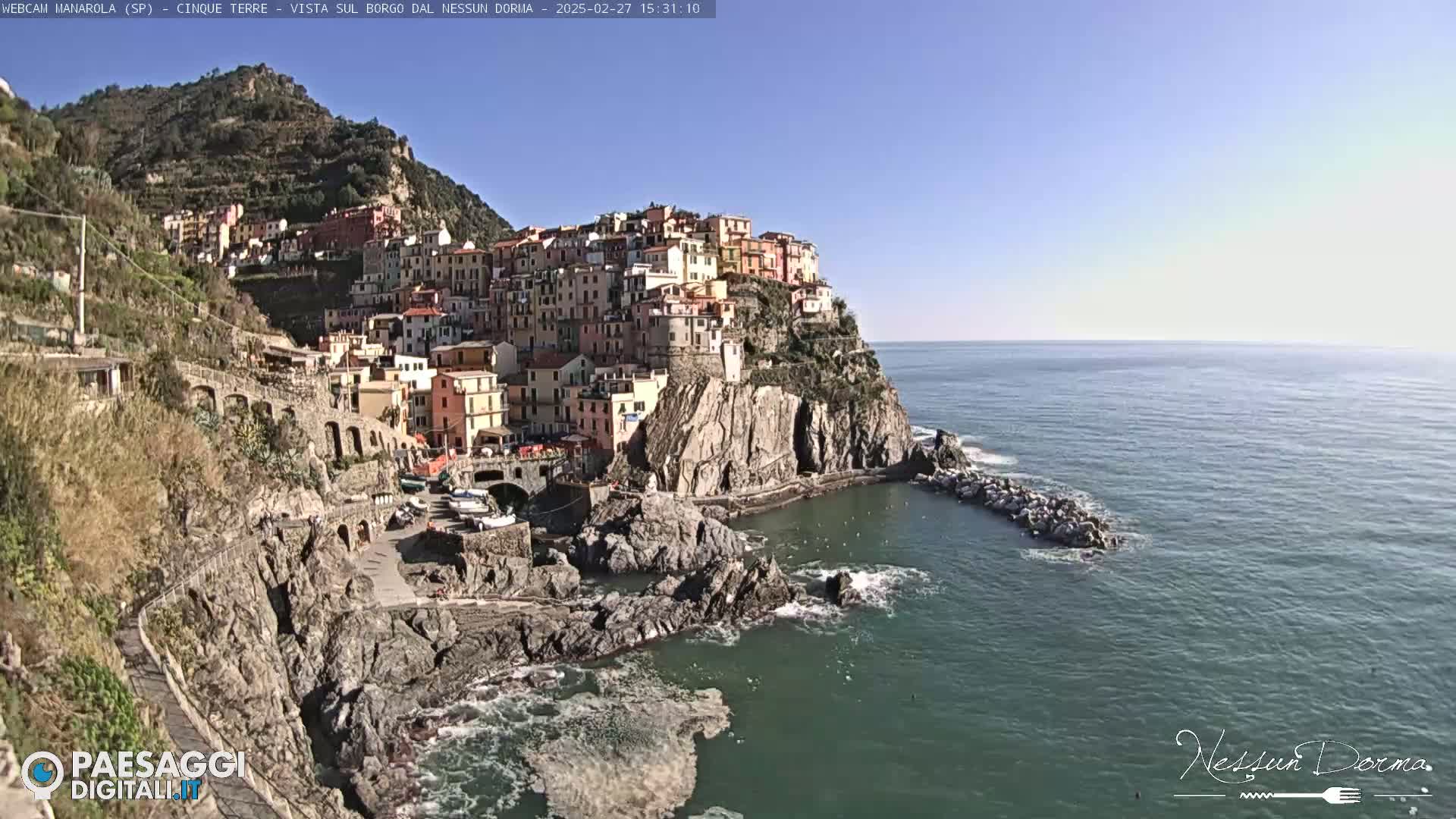 A colorful village clings to a steep cliffside overlooking a calm, blue sea on a sunny day.