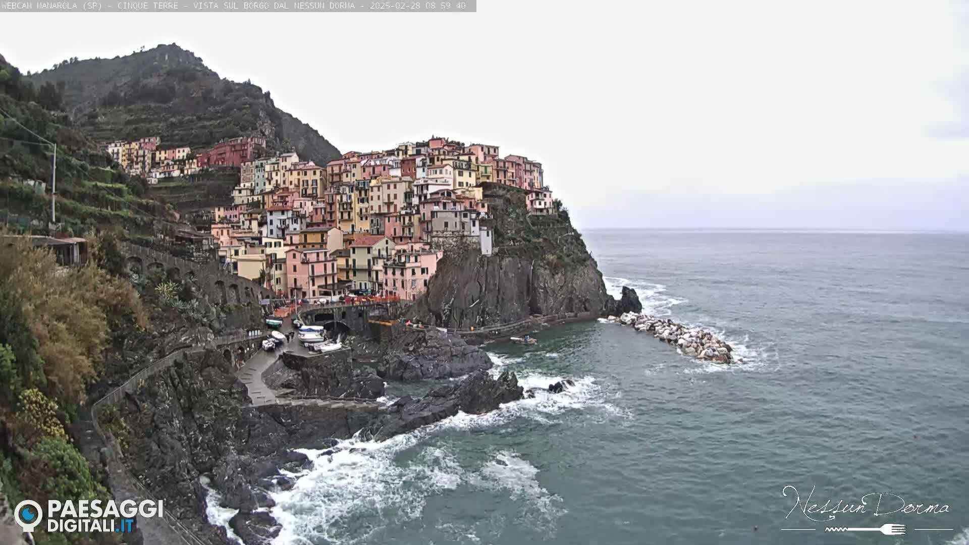 A colorful village clings to a rocky cliffside overlooking a choppy sea under an overcast sky.