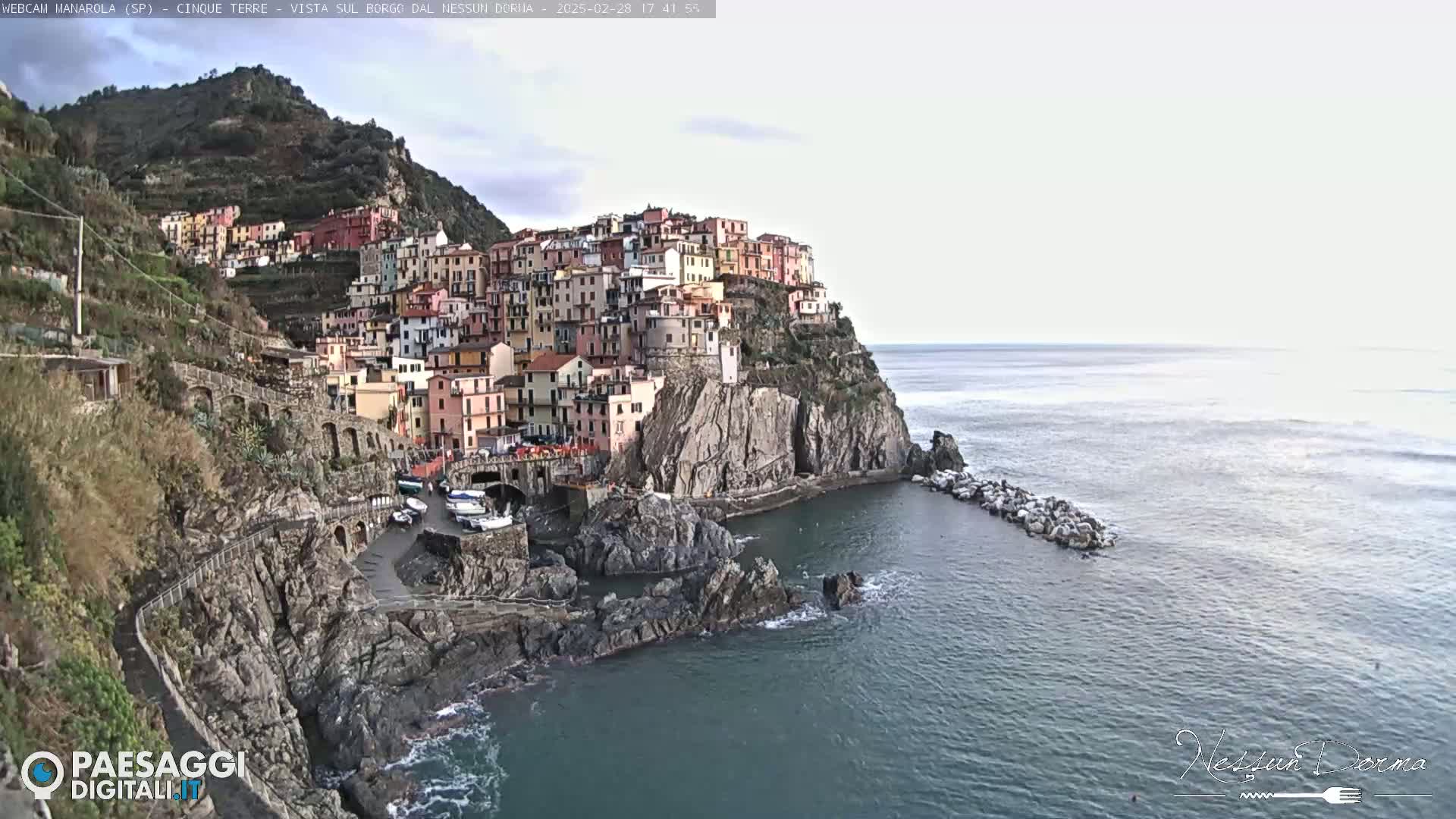 A colorful village clings to a steep cliffside overlooking a calm sea under a mostly cloudy sky.