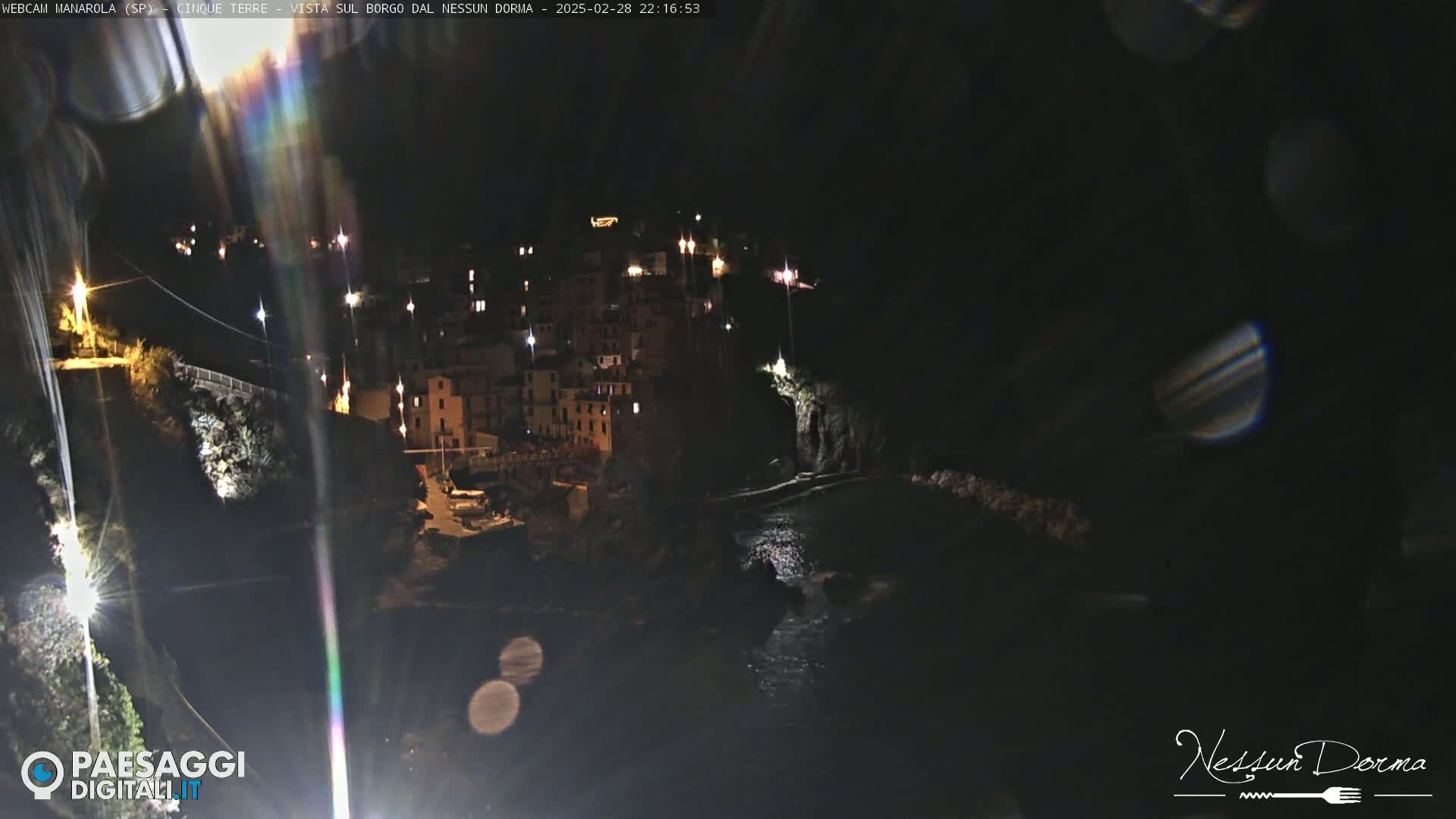 A nighttime view of a coastal town nestled against a dark cliffside, with dimly lit buildings and a calm body of water below.