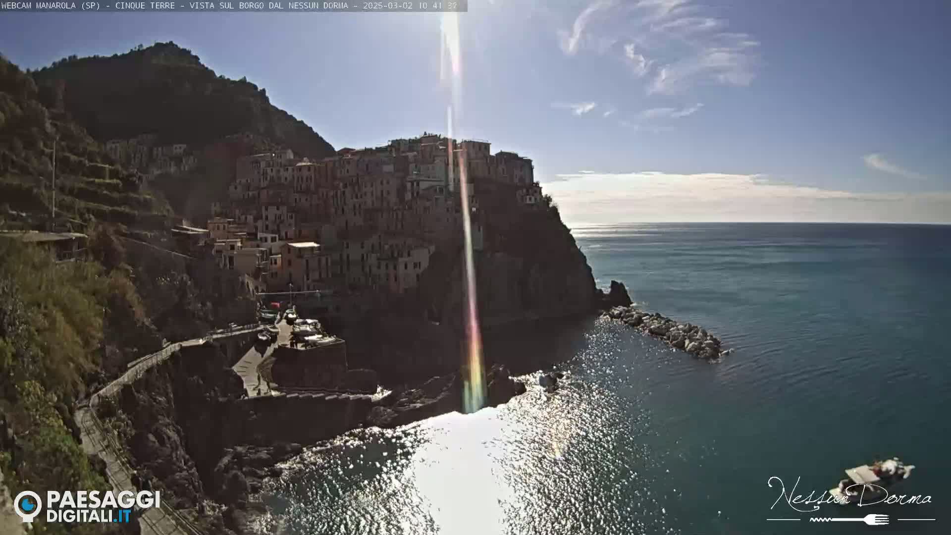 A sunlit coastal village clings to a cliffside overlooking a calm sea on a bright, clear day.