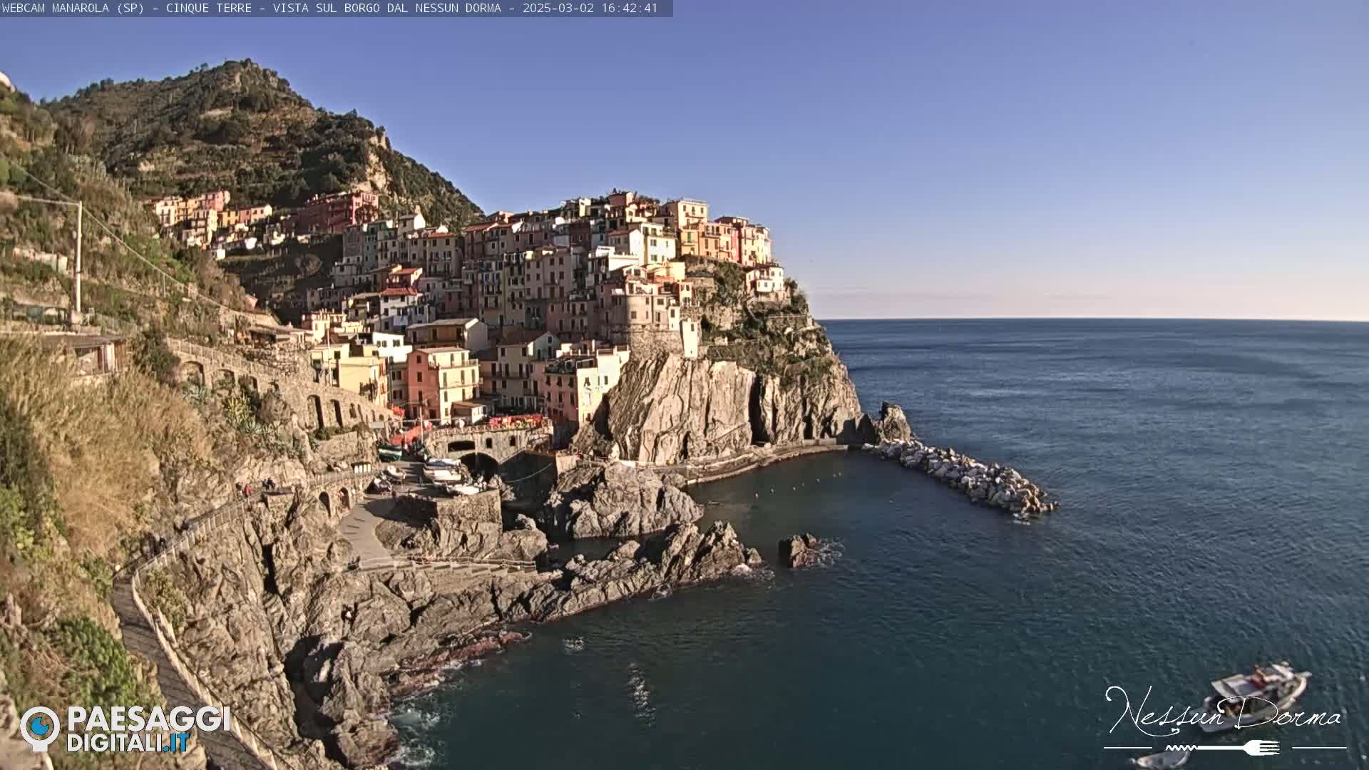 A colorful village clings to a rocky cliffside overlooking a calm blue sea under a clear sky.