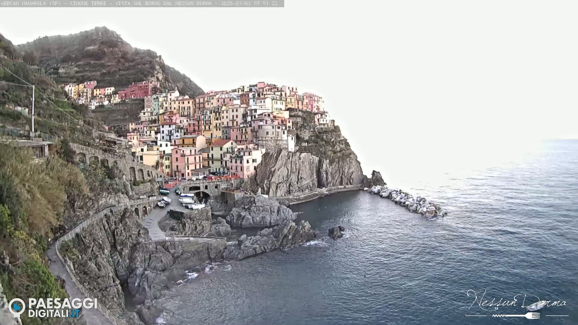 A colorful village clings to a steep cliffside overlooking a calm sea.