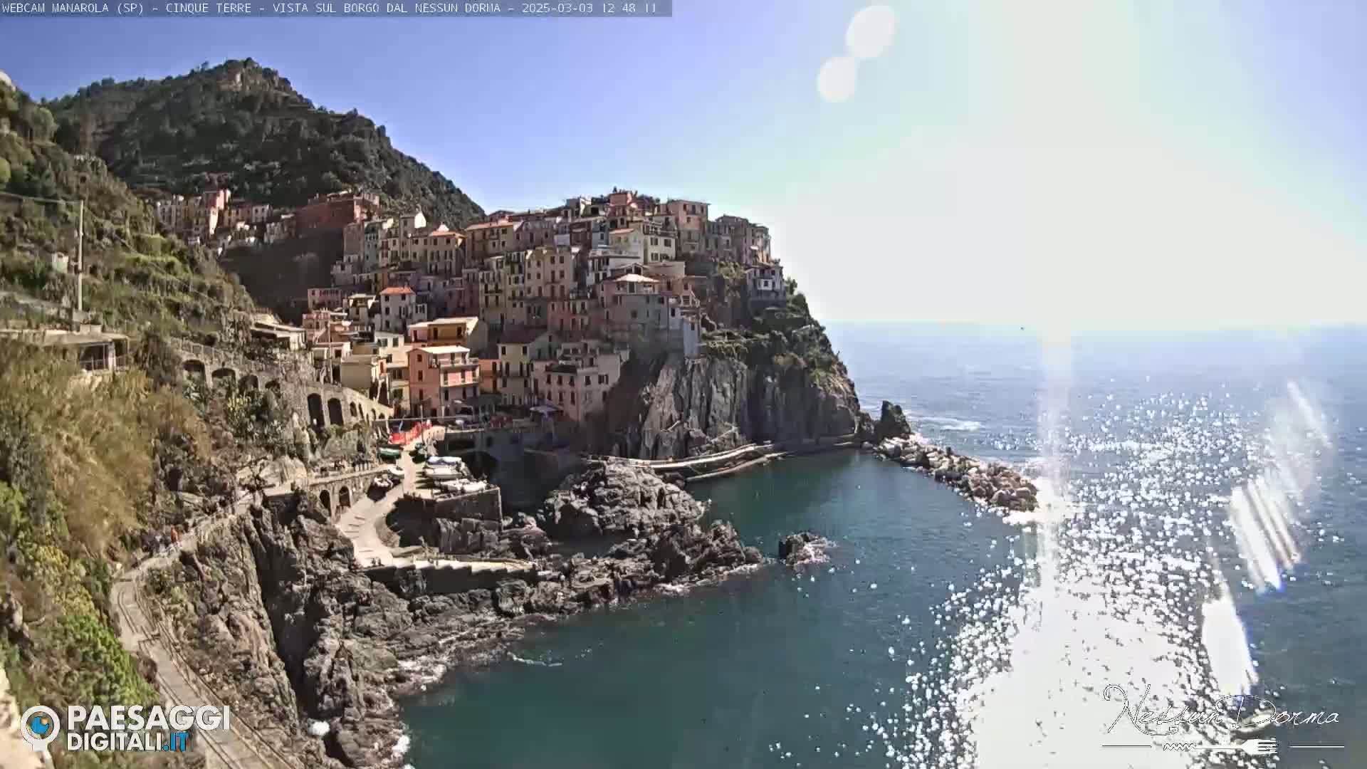 A colorful village clings to a steep cliff overlooking a sparkling blue sea under a bright, sunny sky.