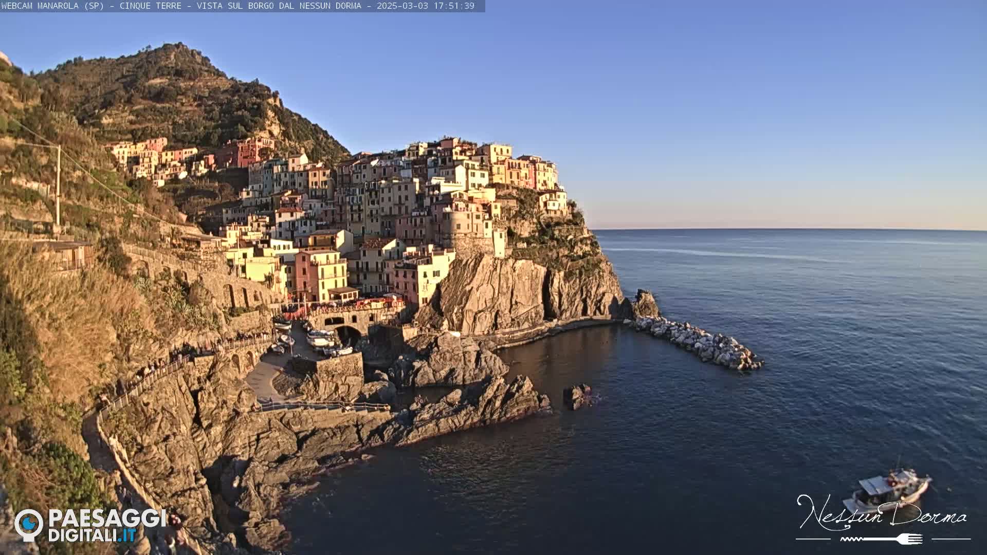 A picturesque coastal town, clinging to a rocky cliffside, is bathed in the warm light of a clear, sunny afternoon, with calm blue waters stretching to the horizon.