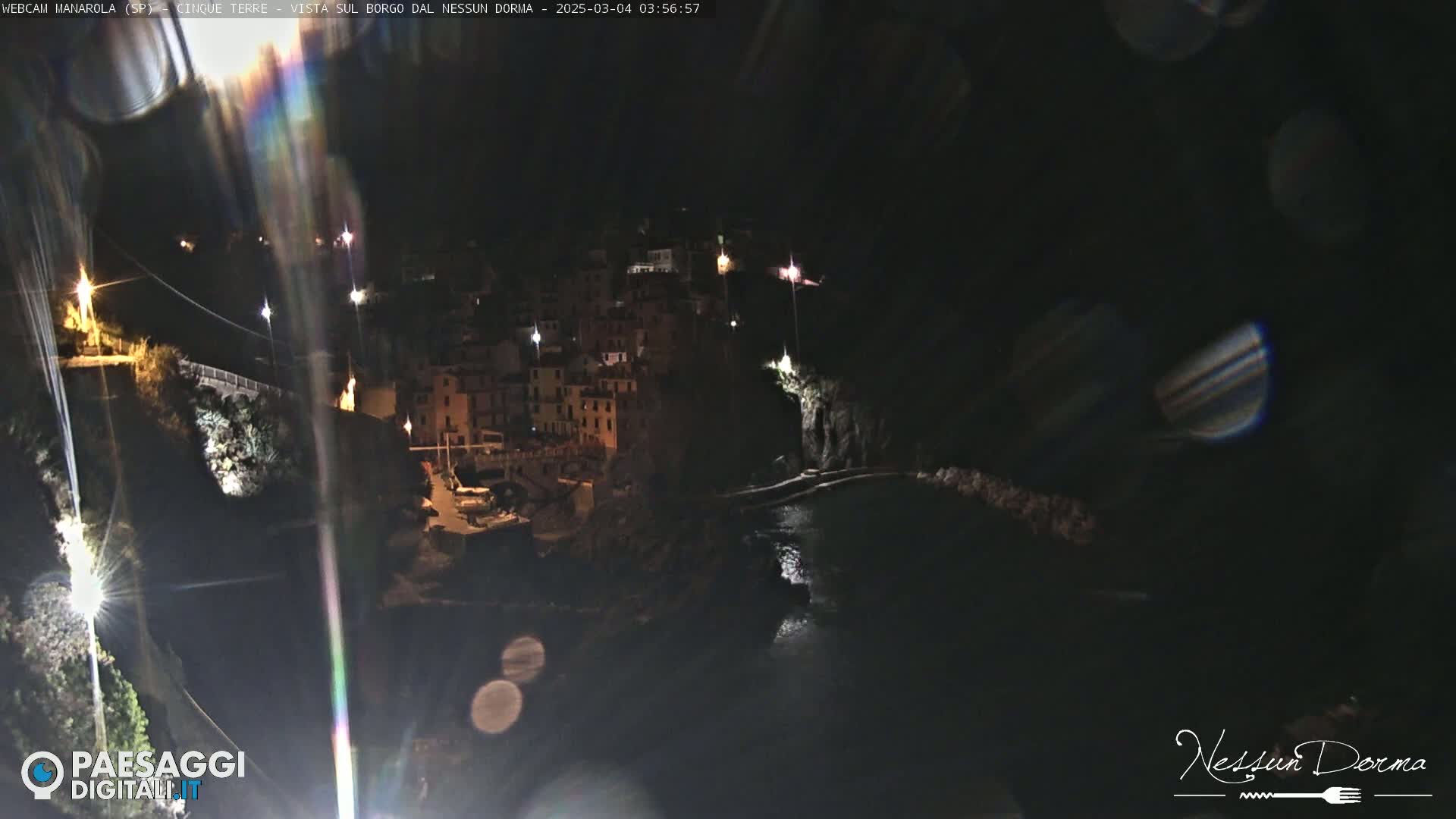 A nighttime, long shot of a coastal village with buildings illuminated along a cliffside overlooking dark water.