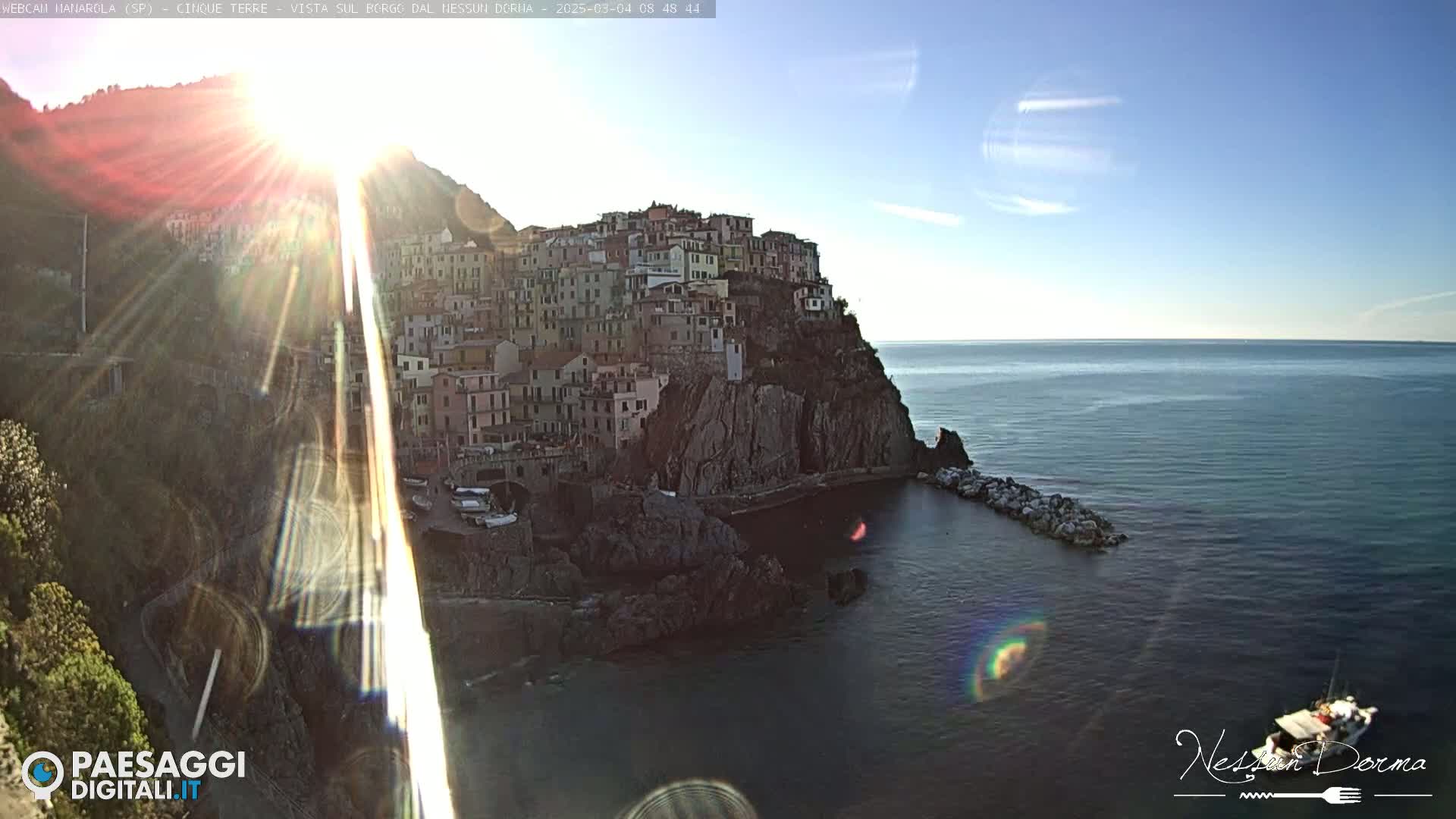 A sunlit coastal town clinging to a rocky cliffside overlooks a calm sea under a mostly clear sky.