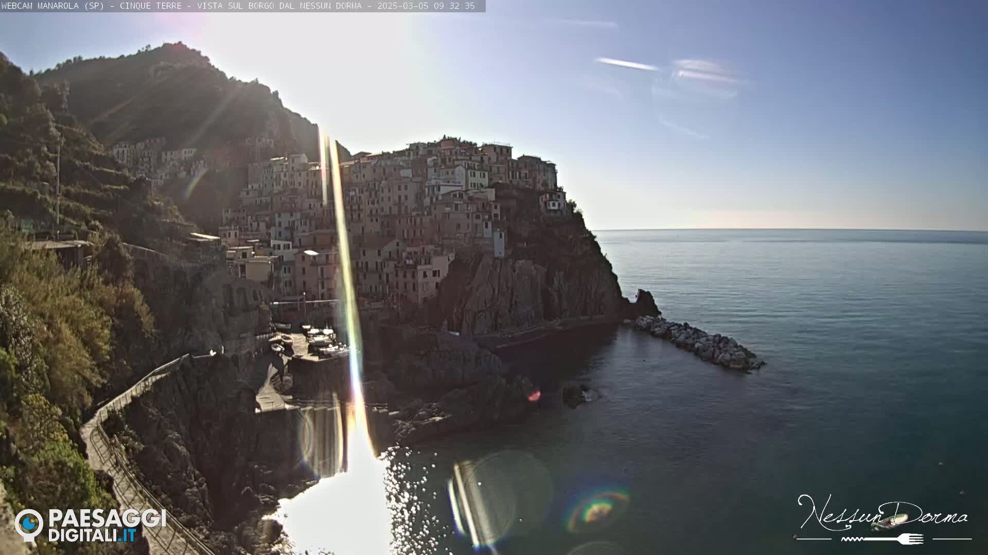 A coastal village clings to a rocky cliffside overlooking a calm sea under a bright, sunny sky.