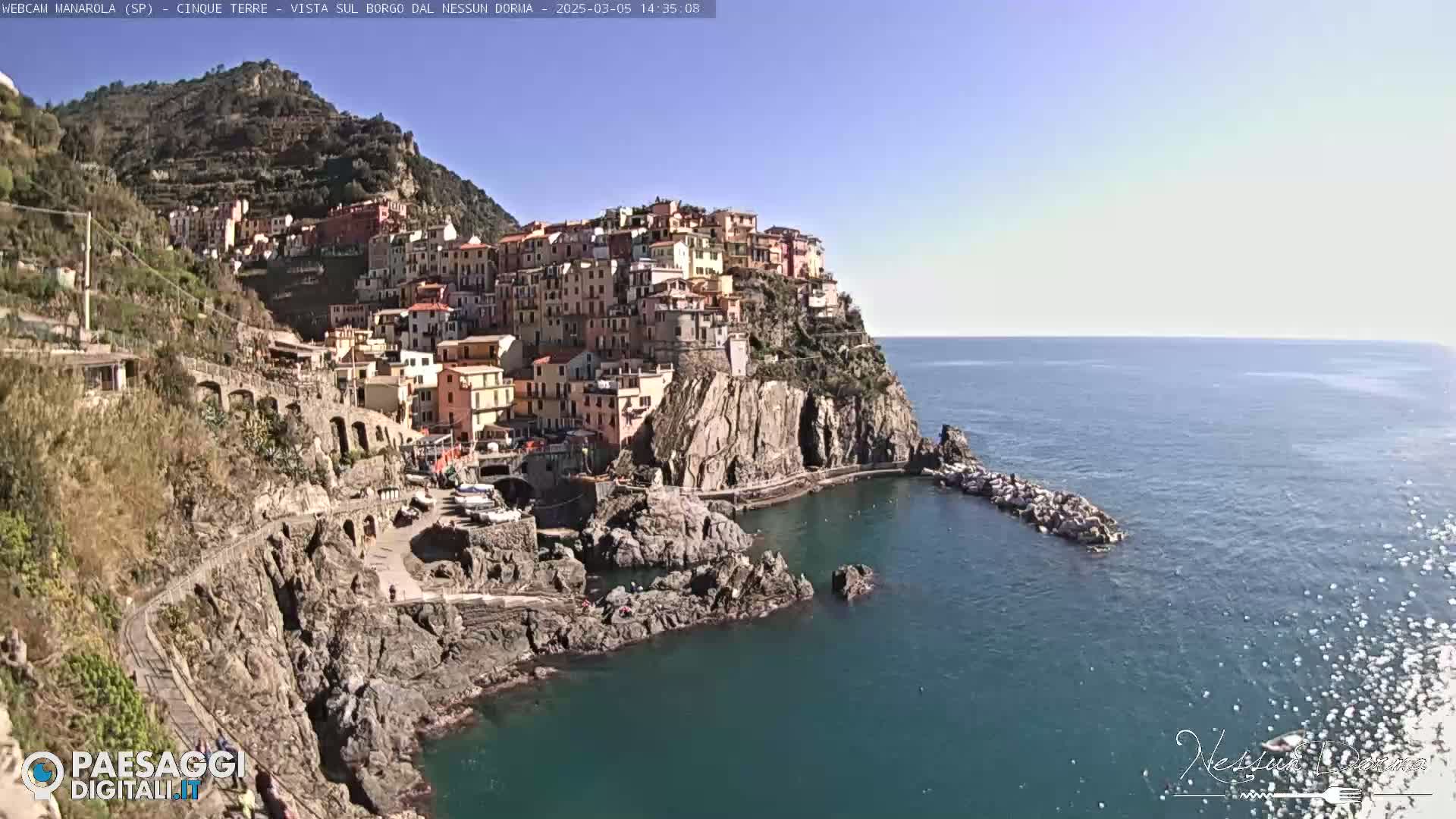 A picturesque coastal town, clinging to a steep cliffside, is nestled beside a calm blue sea under a clear blue sky.