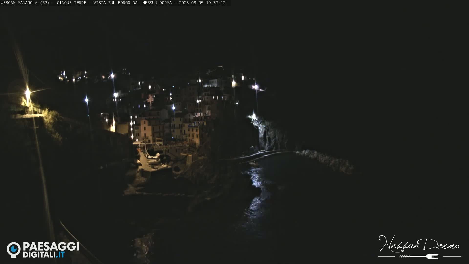 A nighttime, aerial view of a coastal town with buildings illuminated against a dark sky and calm water.