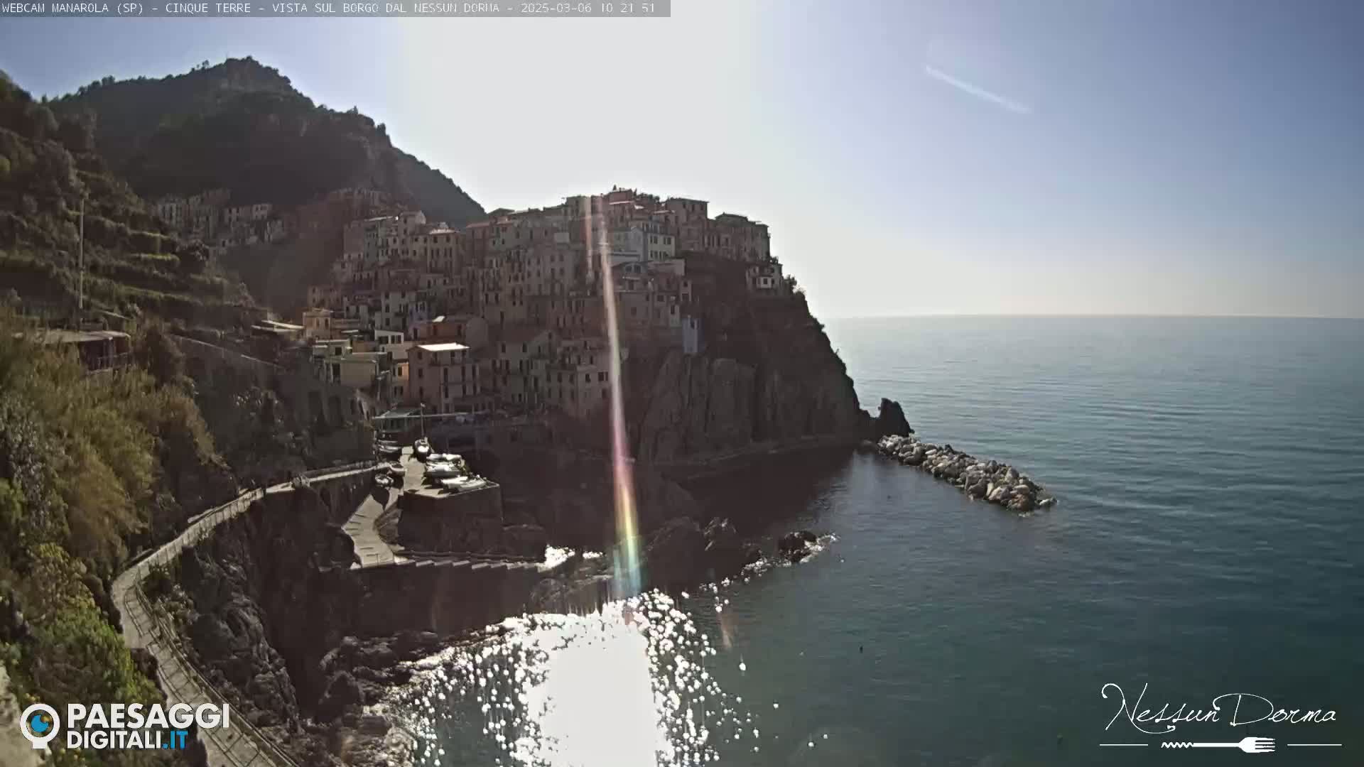 A colorful village clings to a steep cliffside overlooking a calm, sunlit sea.