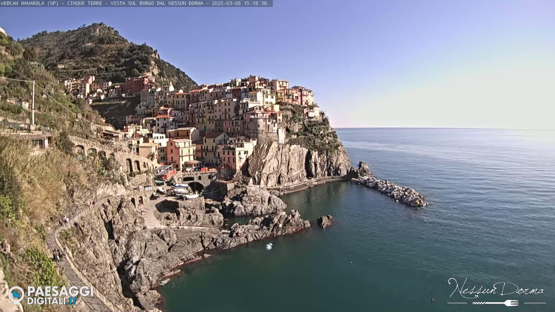 A colorful village clings to a rocky cliffside overlooking a calm, blue sea under a clear, sunny sky.