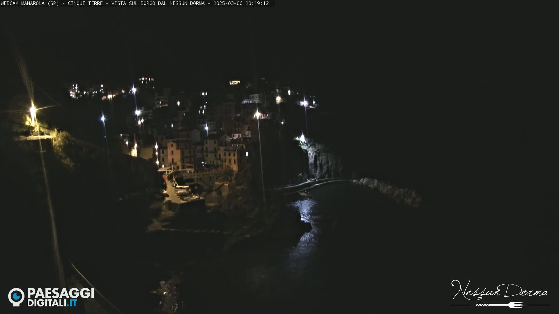A nighttime, aerial view of a coastal town illuminated by lights, with dark water below.
