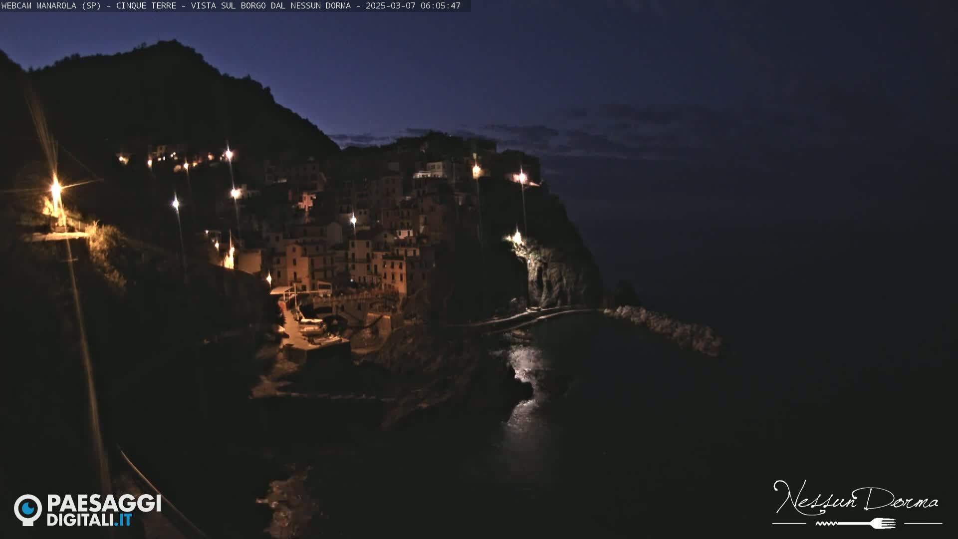 A nighttime, seaside village nestled on a cliffside is illuminated by scattered lights under a dark sky.