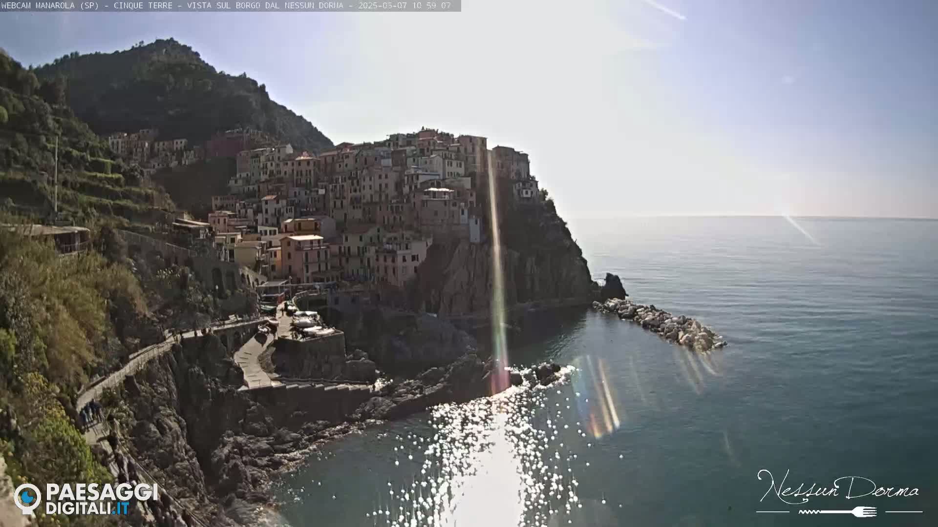 A sunny day reveals a colorful village clinging to a steep cliffside overlooking a calm sea.