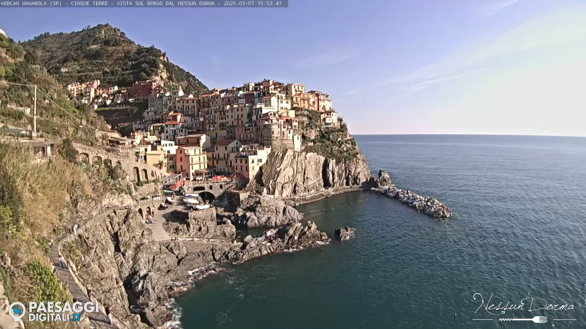 A picturesque coastal village clings to a steep cliffside overlooking a calm blue sea under a clear sky.
