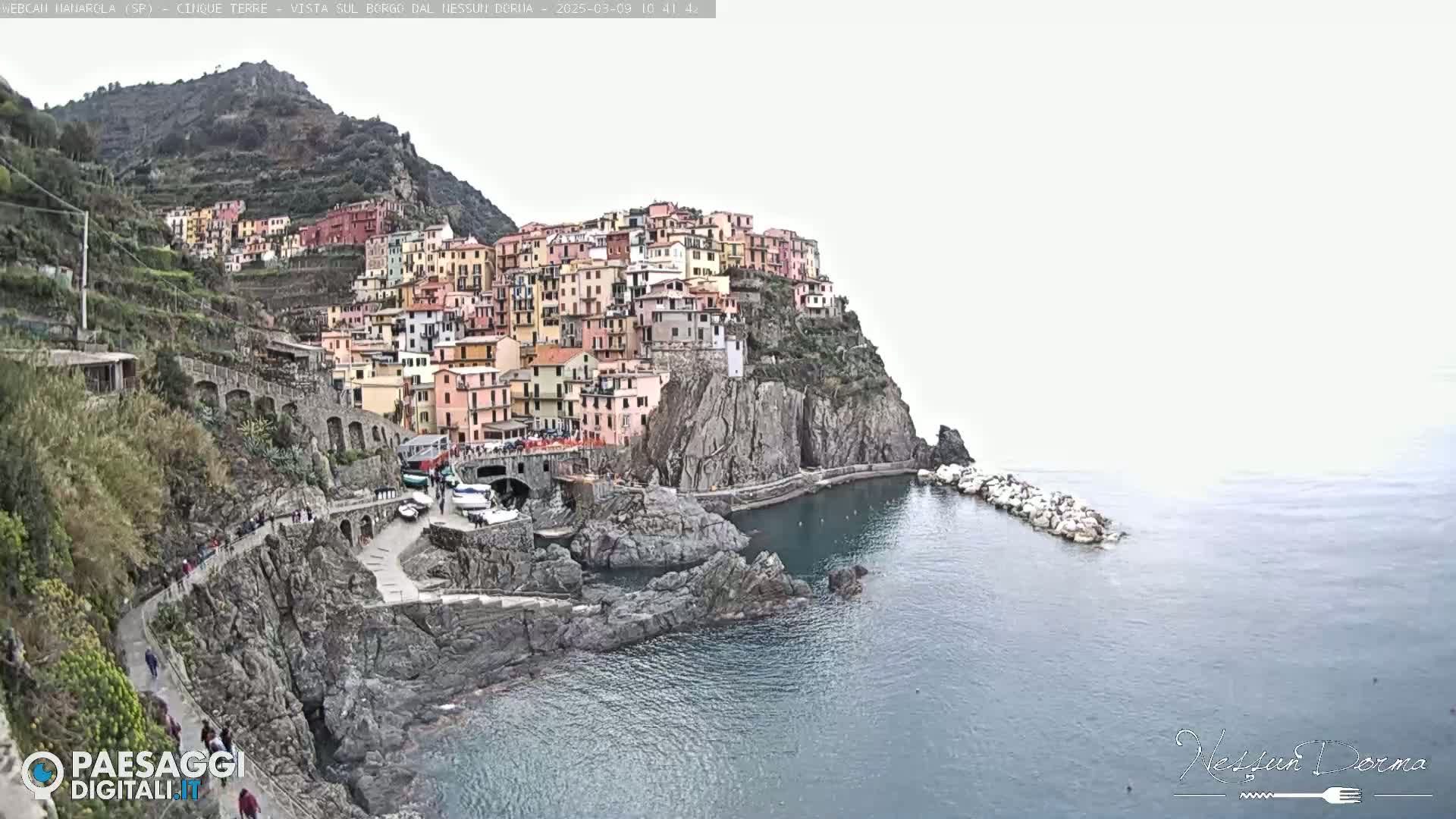 A colorful village clings to a steep cliffside overlooking a calm sea on an overcast day.