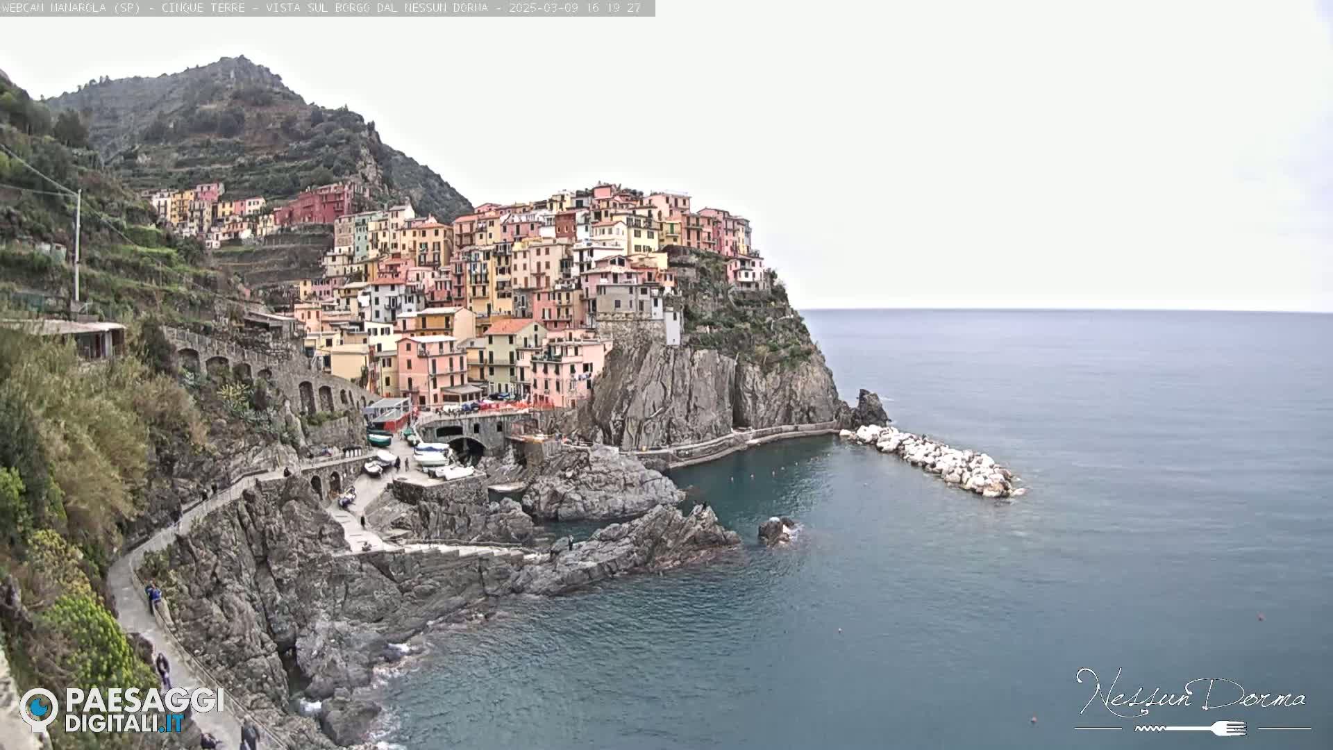 A colorful village clings to a steep cliffside overlooking a calm sea under an overcast sky.