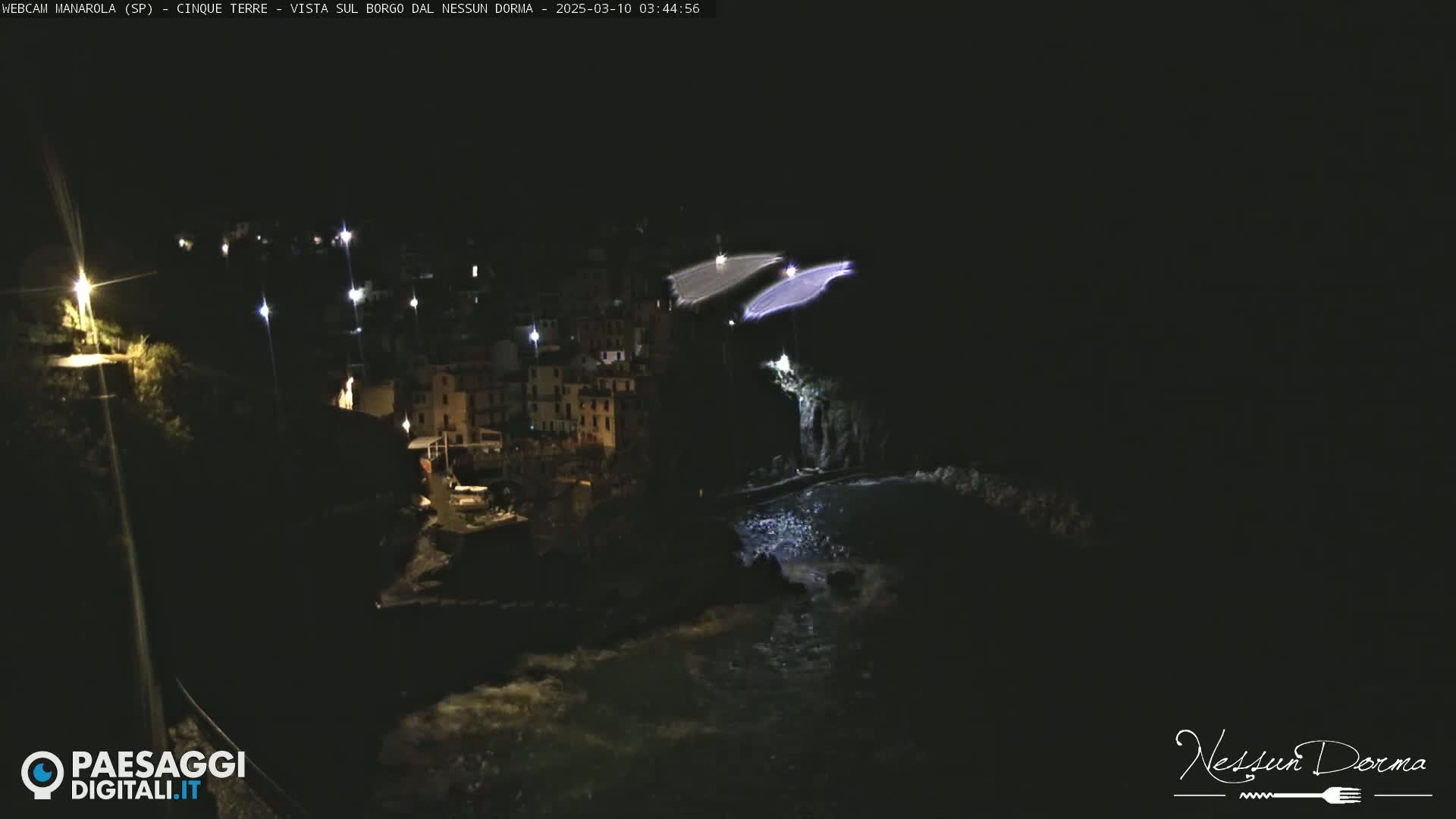 A nighttime coastal village is illuminated, with dark water lapping at the shore.