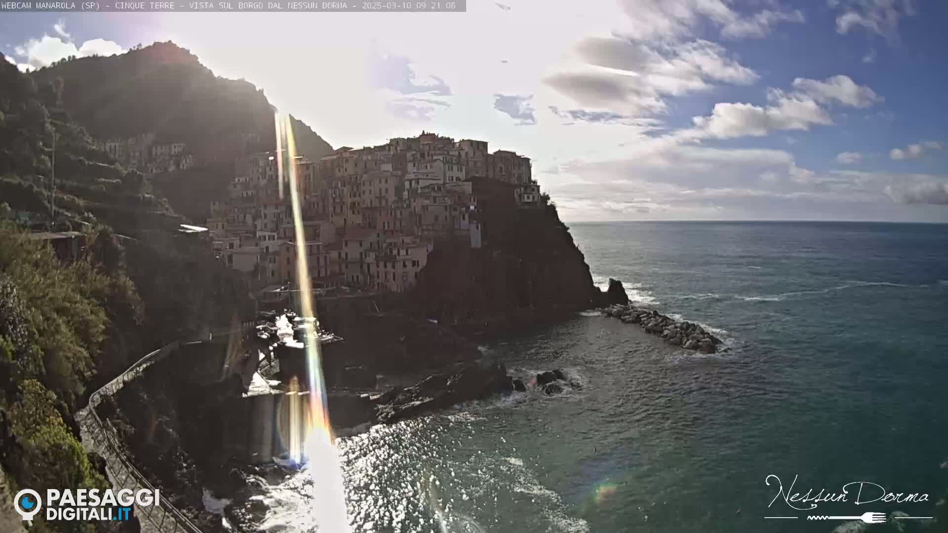 A sunlit coastal town built onto a cliffside overlooking a calm sea under a partly cloudy sky.